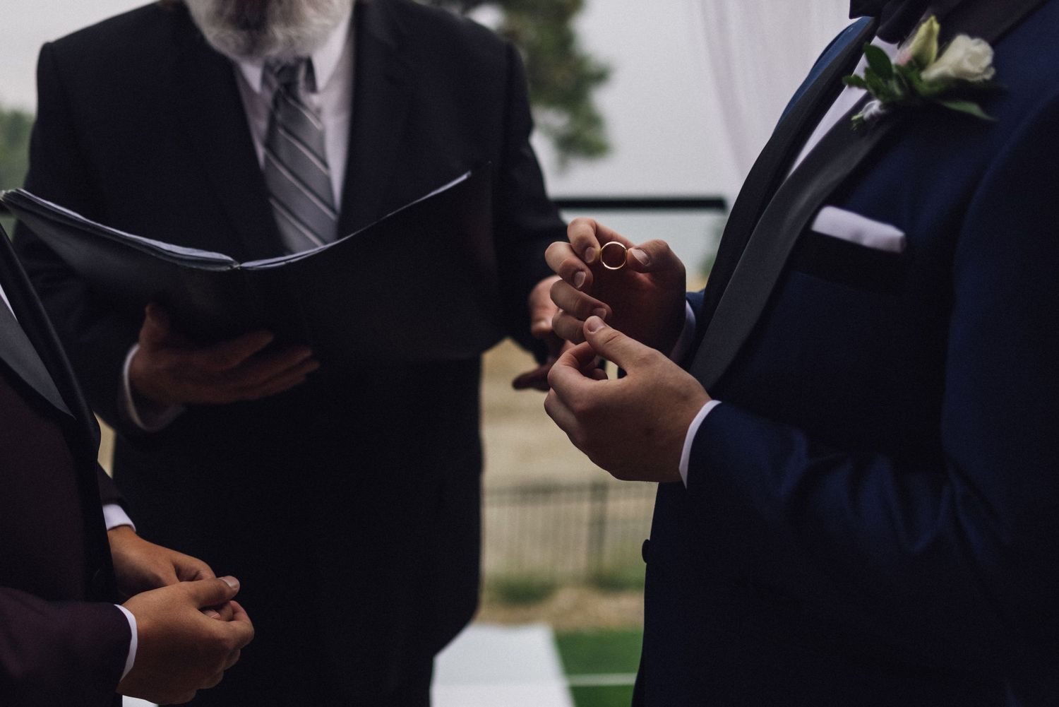 Groom placing wedding ring on partner's finger during outdoor ceremony, officiant holding black book in background.
