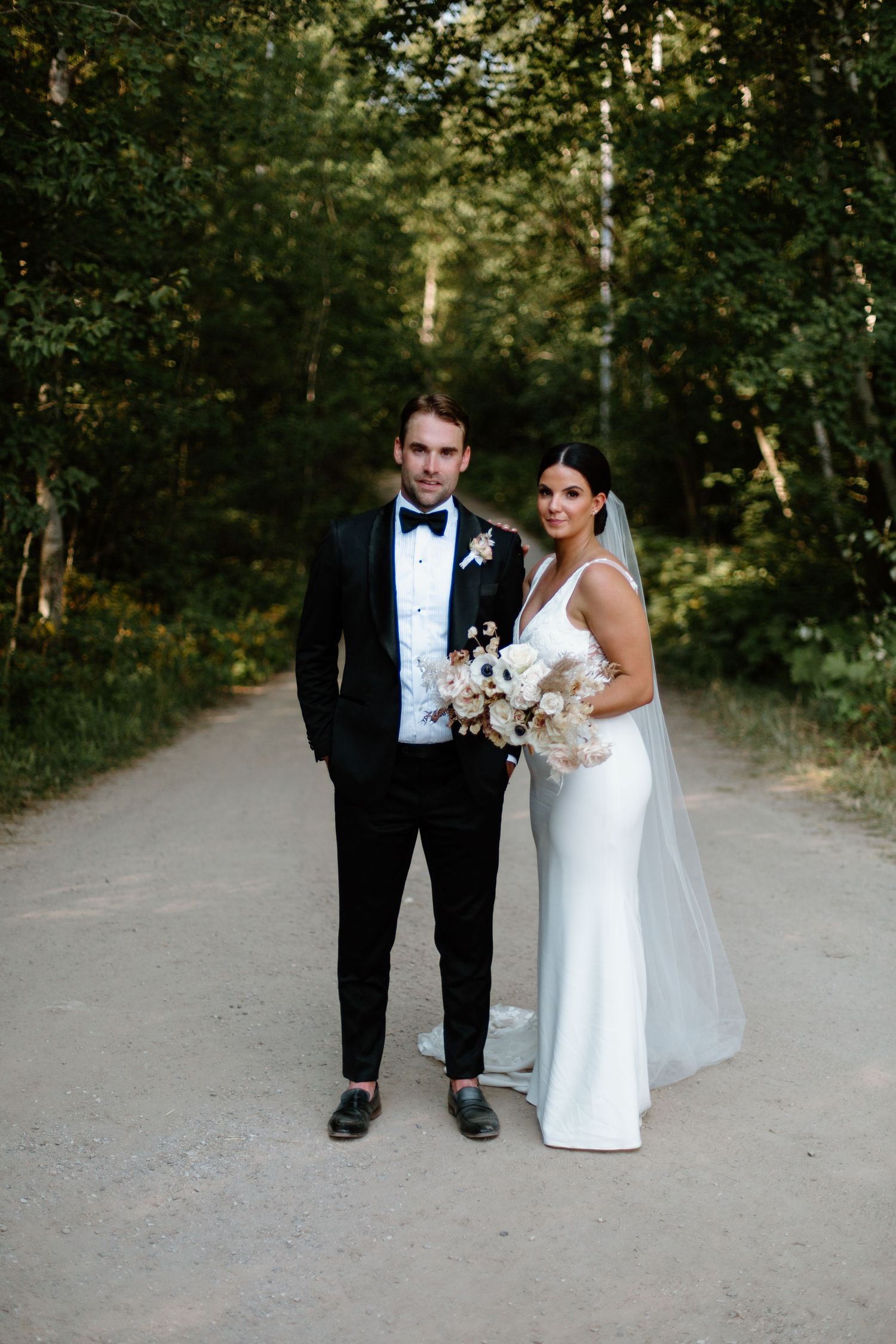 A wedding couple poses on a tree-lined path in formal attire with a white bouquet.