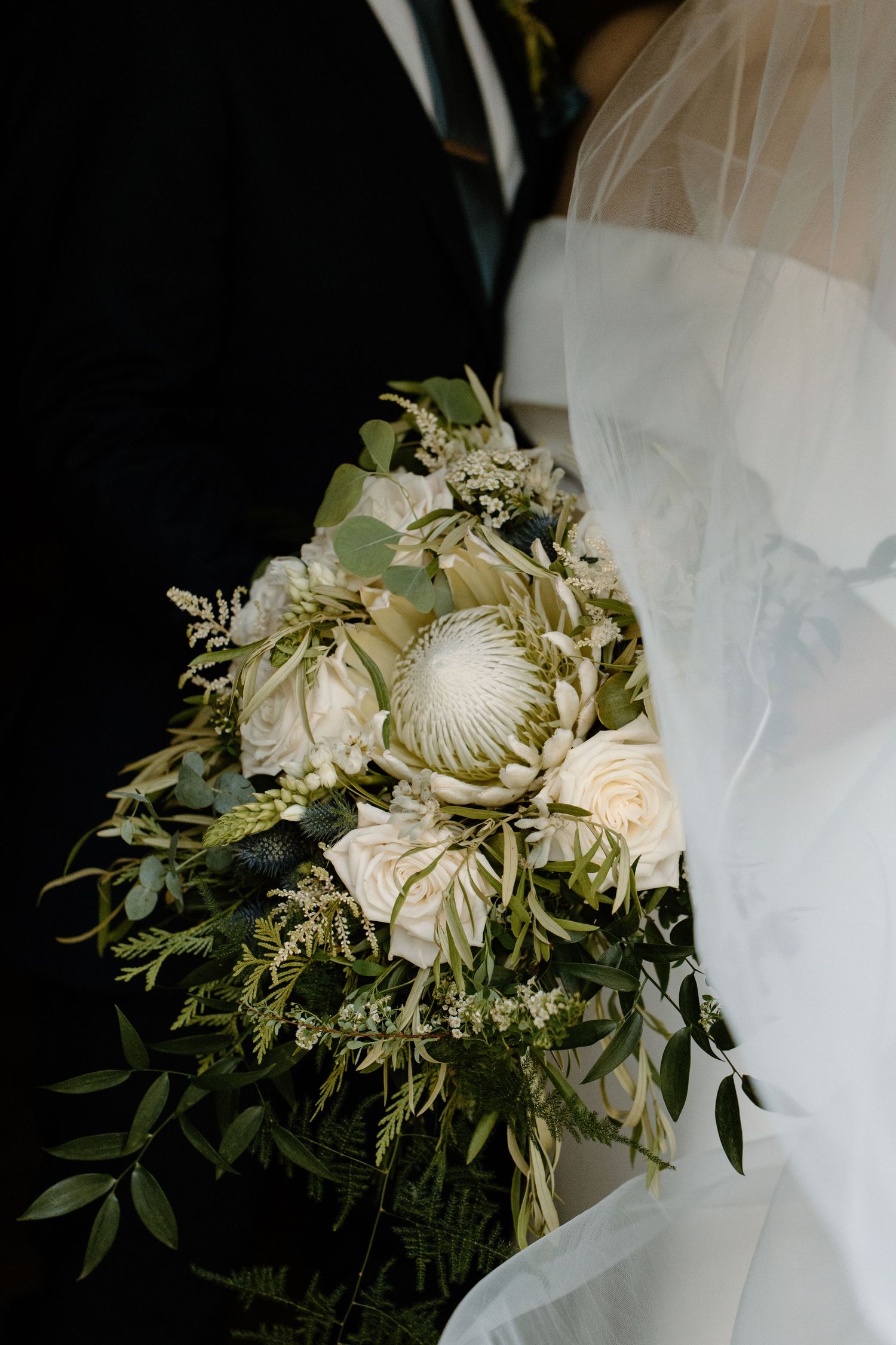 A romantic wedding bouquet with white protea flowers, roses, and delicate greenery against a dark background.