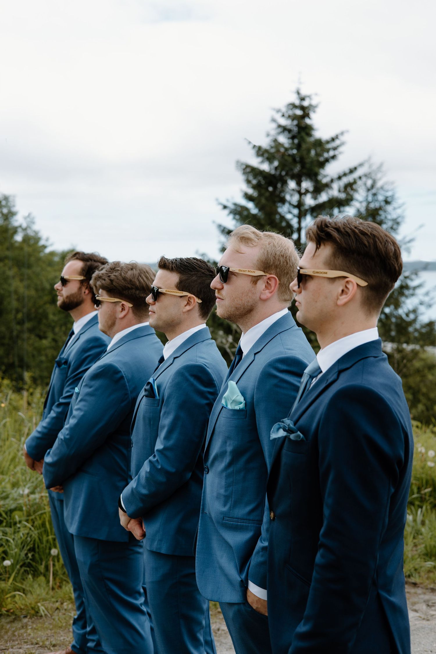 Five groomsmen in matching blue suits and sunglasses stand in a line outdoors at a wedding ceremony.