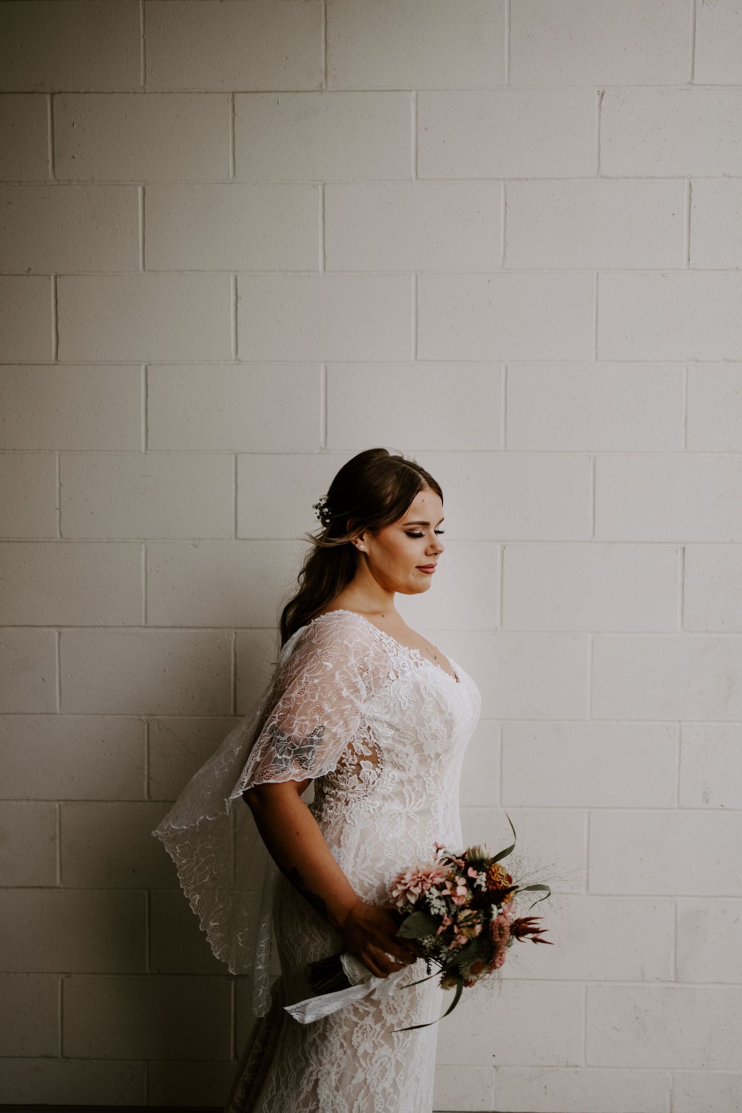 Wedding dress portrait against white brick wall with delicate lace details and bouquet.