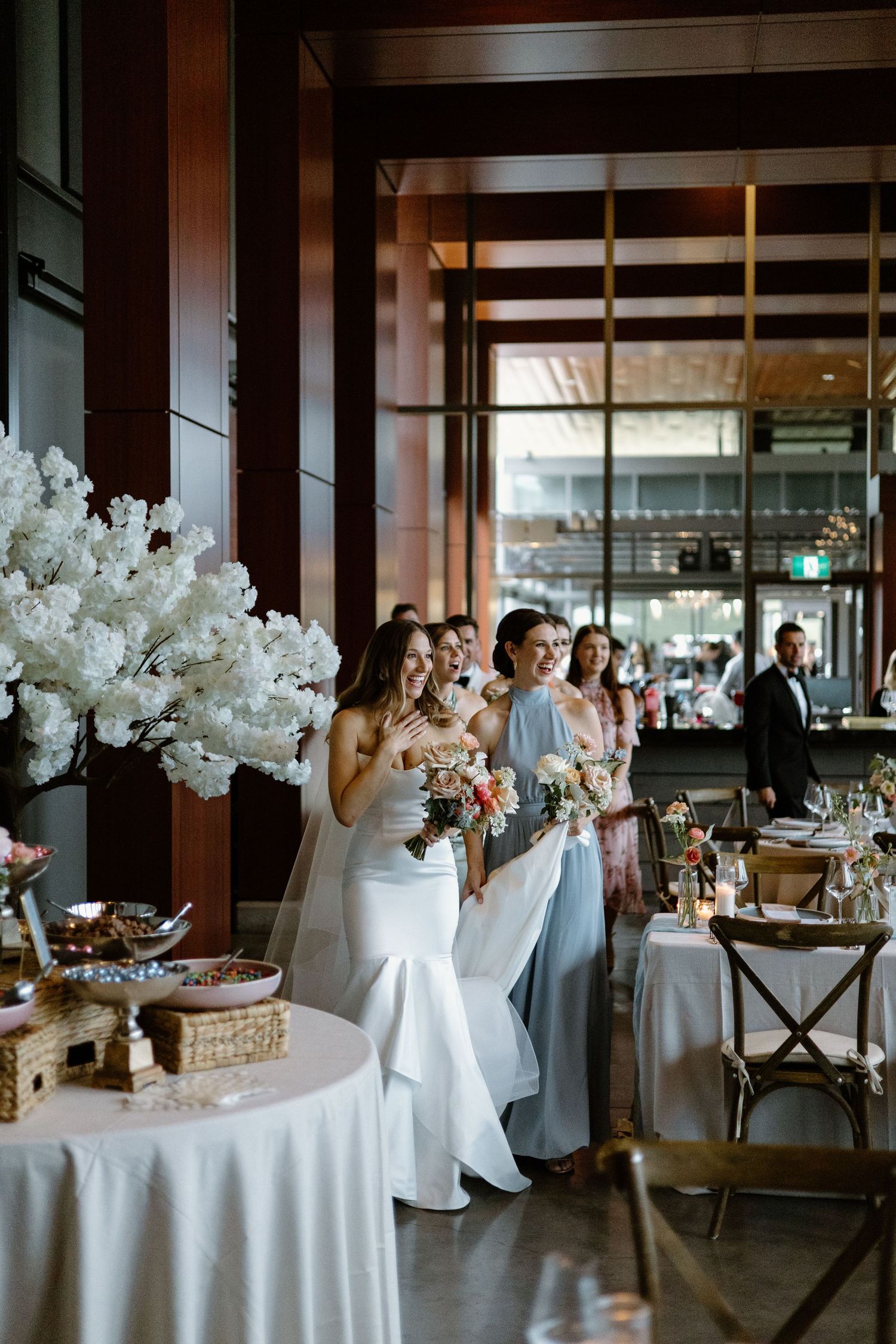 Modern wedding reception venue with white tablecloths, floral centerpieces and wooden cross-back chairs.