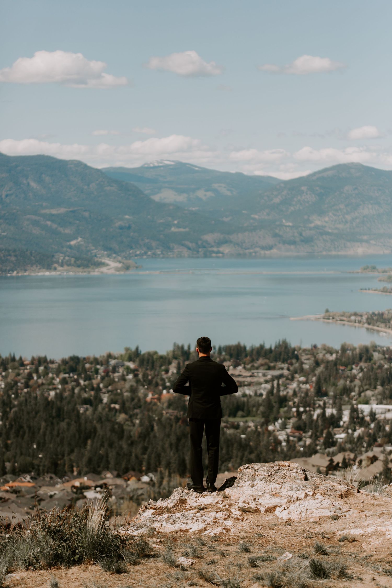 Person standing on mountain overlook gazing at scenic lake and mountain landscape with town below.