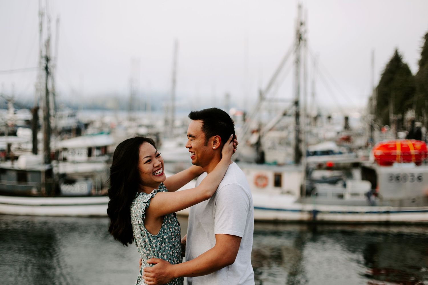 Couple embracing by a marina with sailboats and overcast skies in the background.