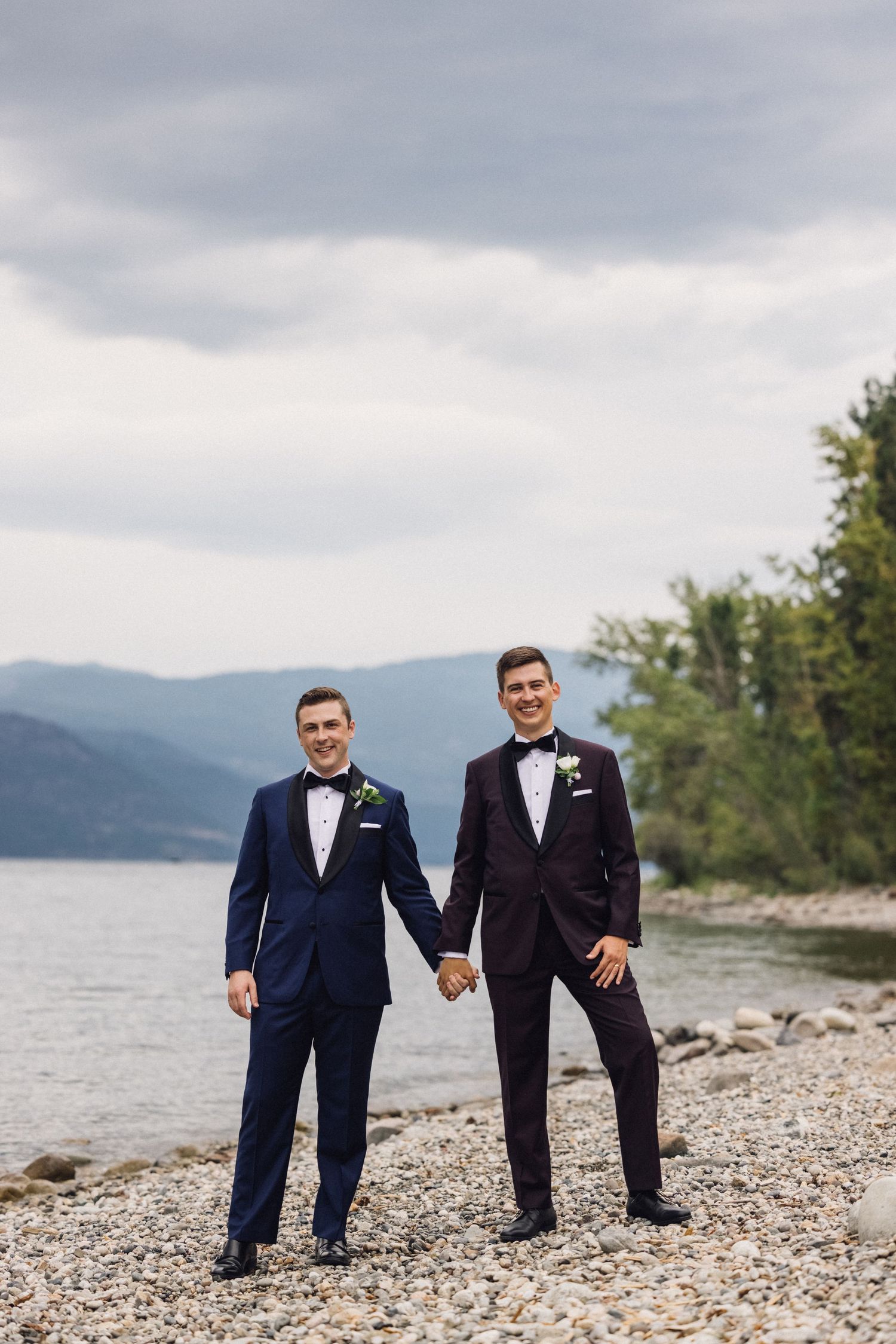 Wedding couple and groomsmen pose creatively on a rocky beach shore with mountains and lake in the background.