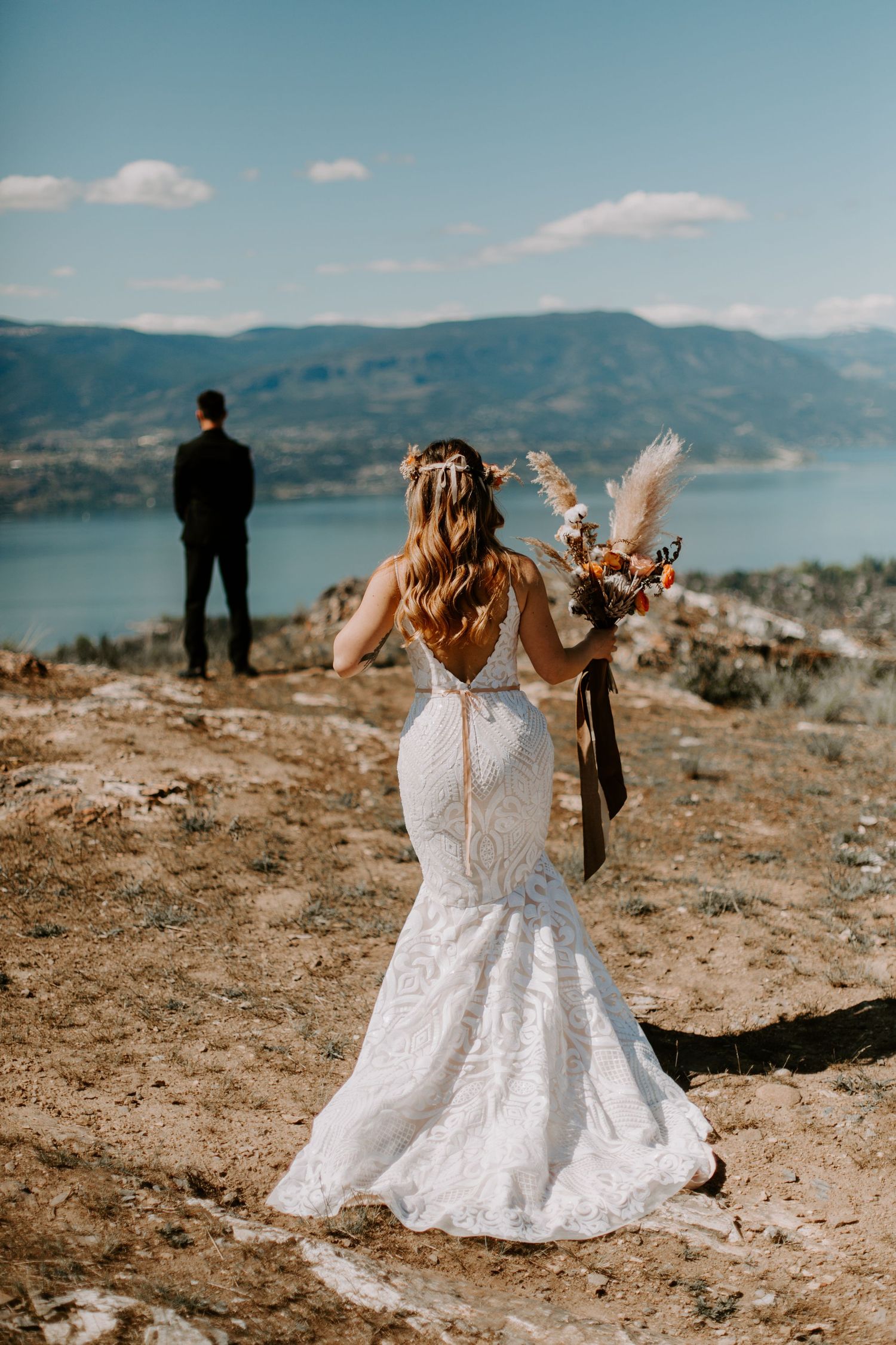 Two people in white wedding dresses sit together on a rocky overlook with mountain and lake views.