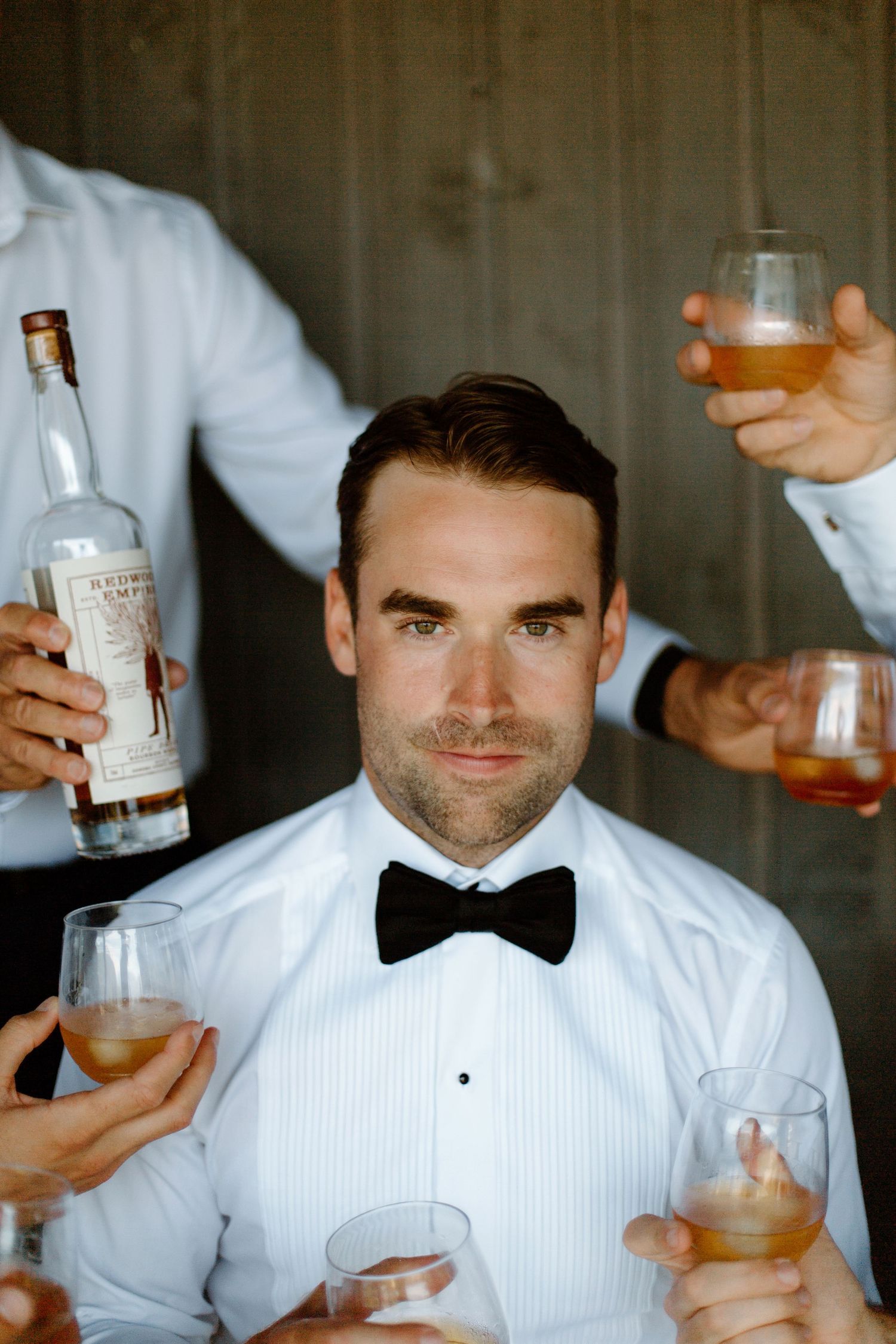 A person in a white tuxedo shirt and bow tie smiles while holding a drink at what appears to be a wedding reception.