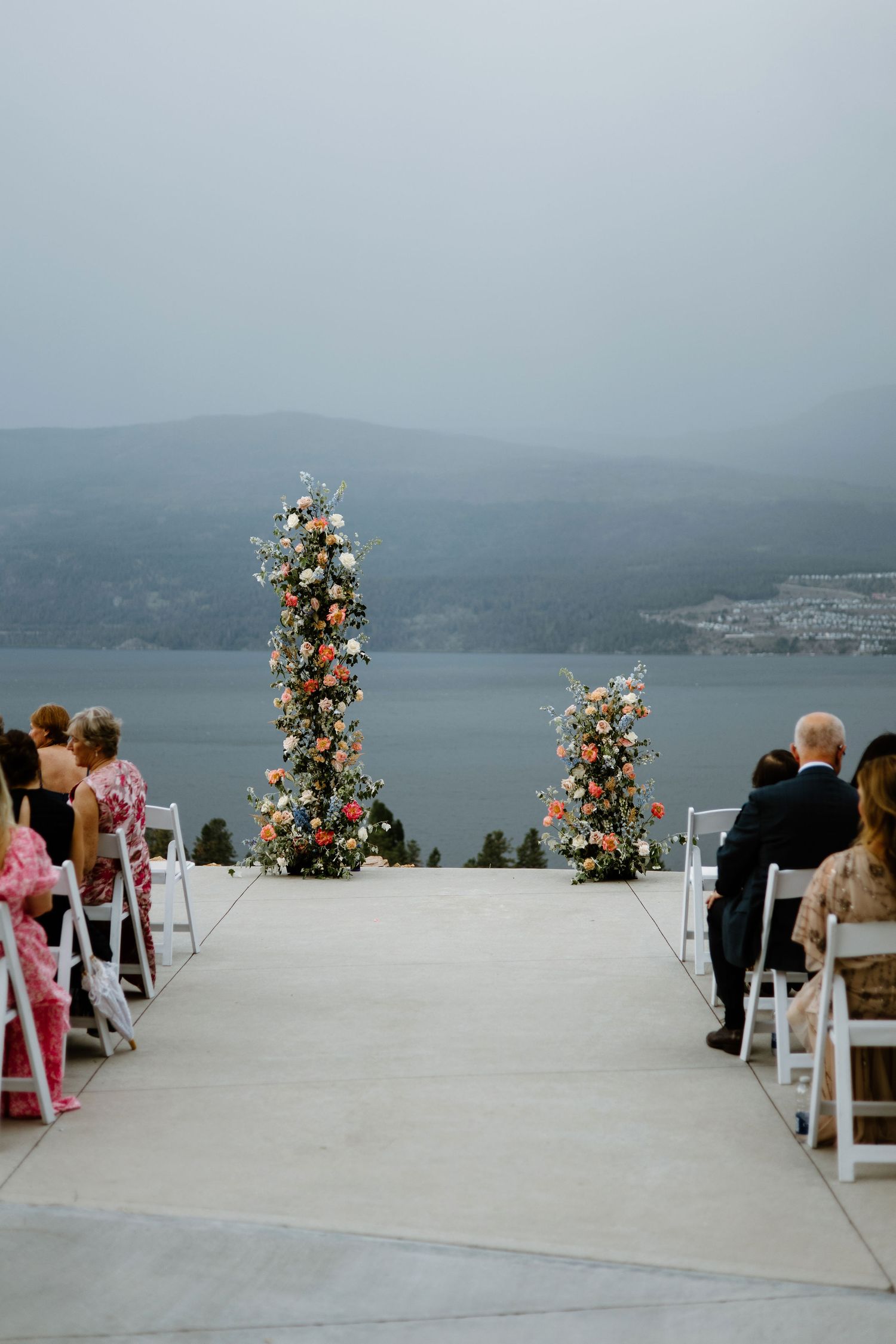 Outdoor wedding ceremony on a terrace overlooking a misty lake, with tall floral columns and guests seated in white chairs.