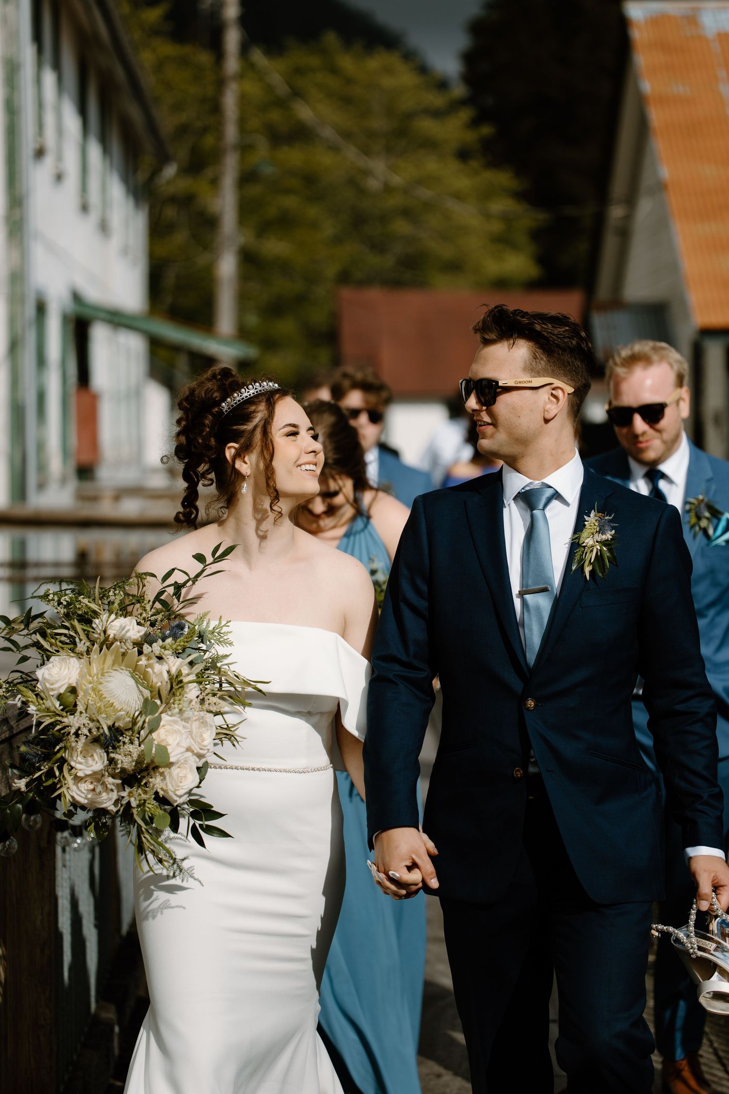 A wedding couple walks together on a sunny day with the wedding party following behind in navy blue attire.