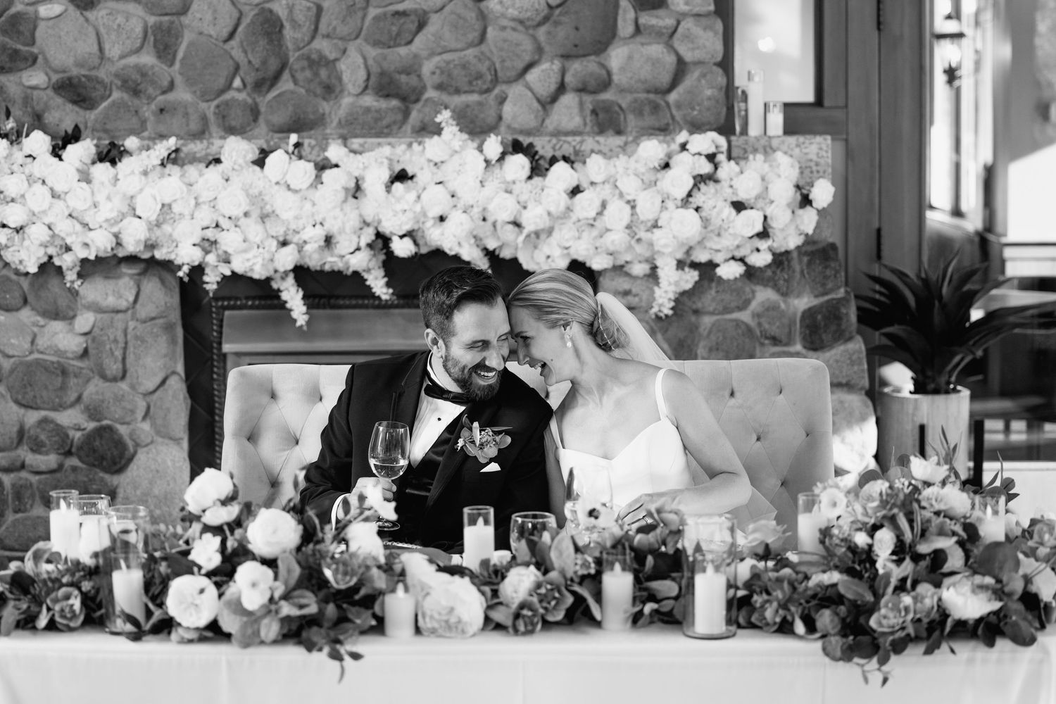 A black and white wedding photo showing a couple sharing a joyful moment at their reception table with floral decorations.