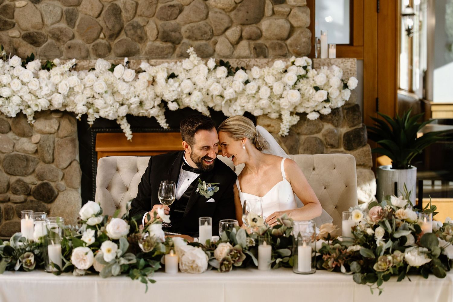 Bride and groom share an intimate moment at floral sweetheart table with white roses and candles at elegant wedding reception.
