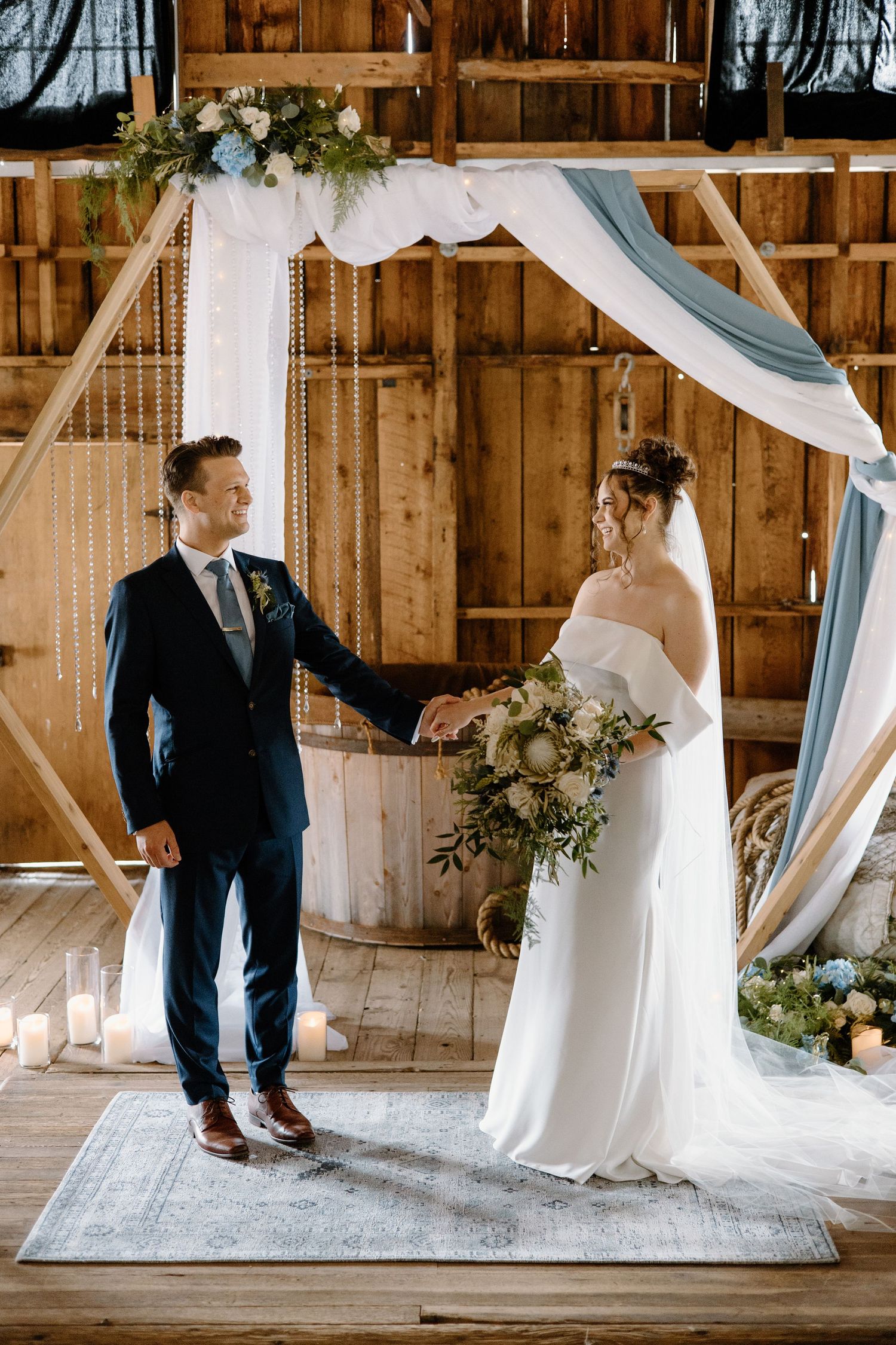 Bride and groom hold hands at rustic barn wedding ceremony under geometric wooden arch with blue draping and floral decor.