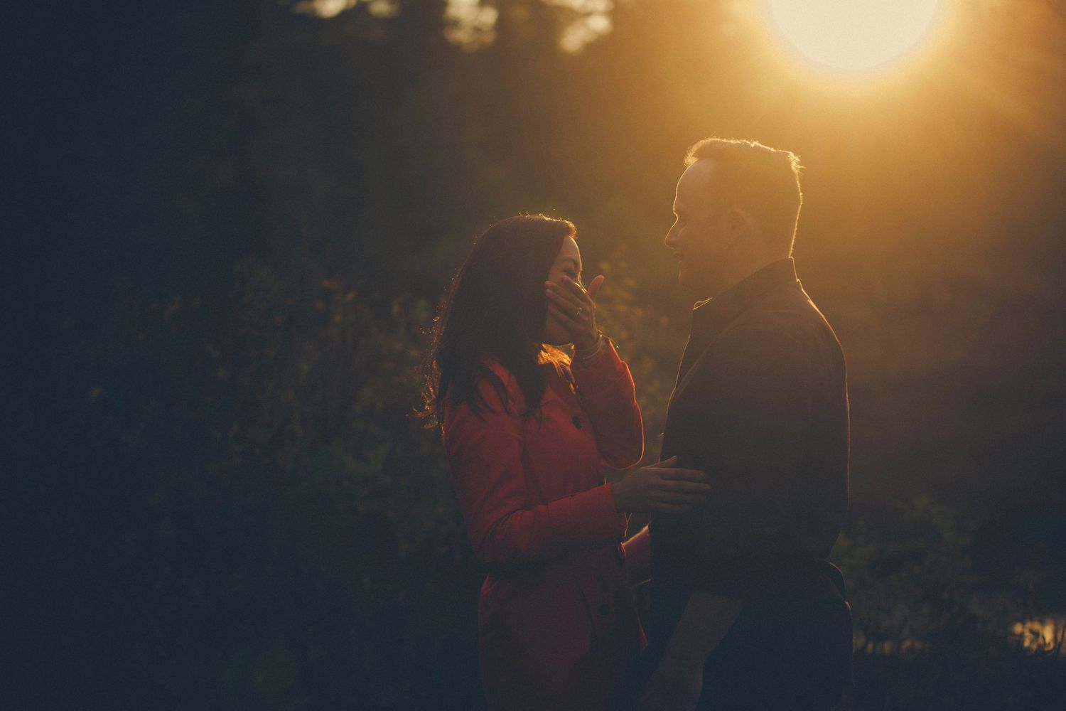 Couple sharing a romantic moment at golden sunset in a forest, warm backlit glow highlighting their embrace.