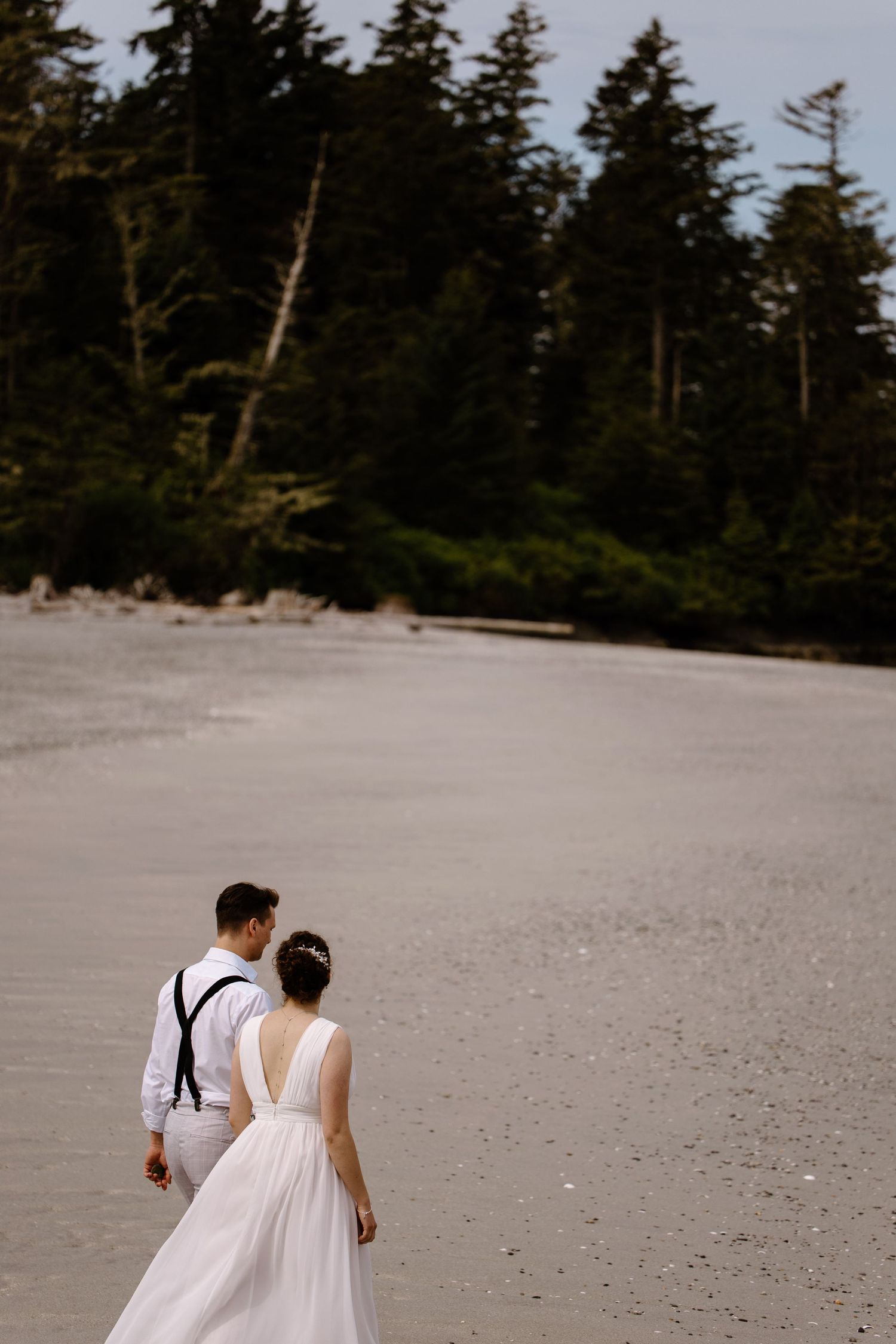 Couple in white walking along a serene beach with forested coastline.