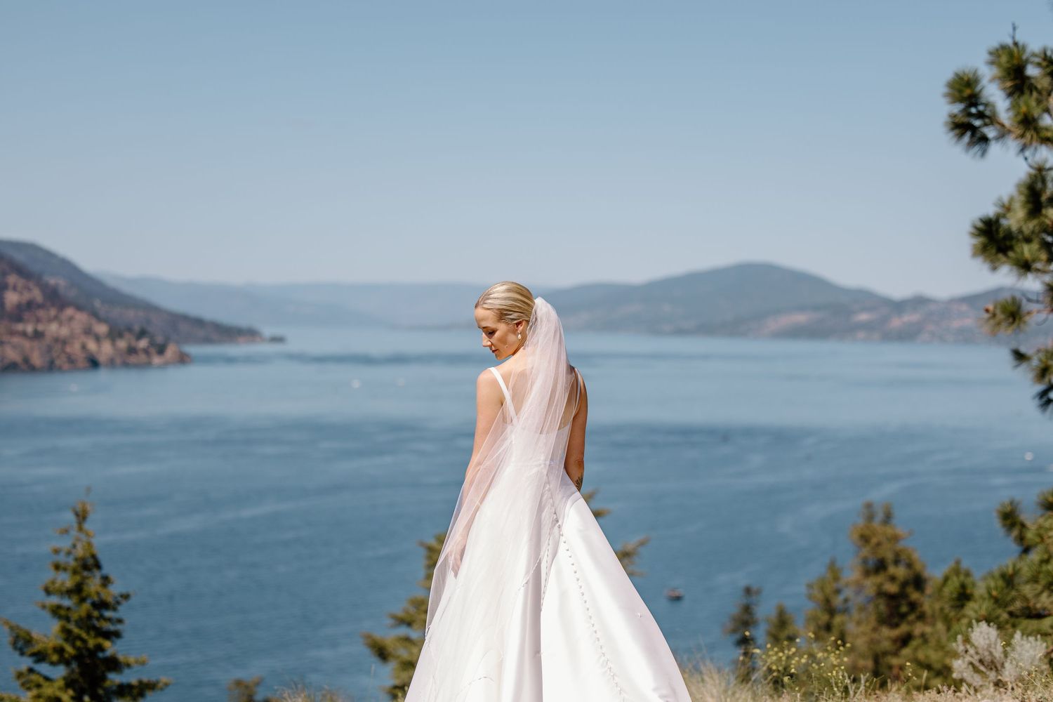 A wedding dress flows elegantly against a scenic mountain lake backdrop on a sunny day.