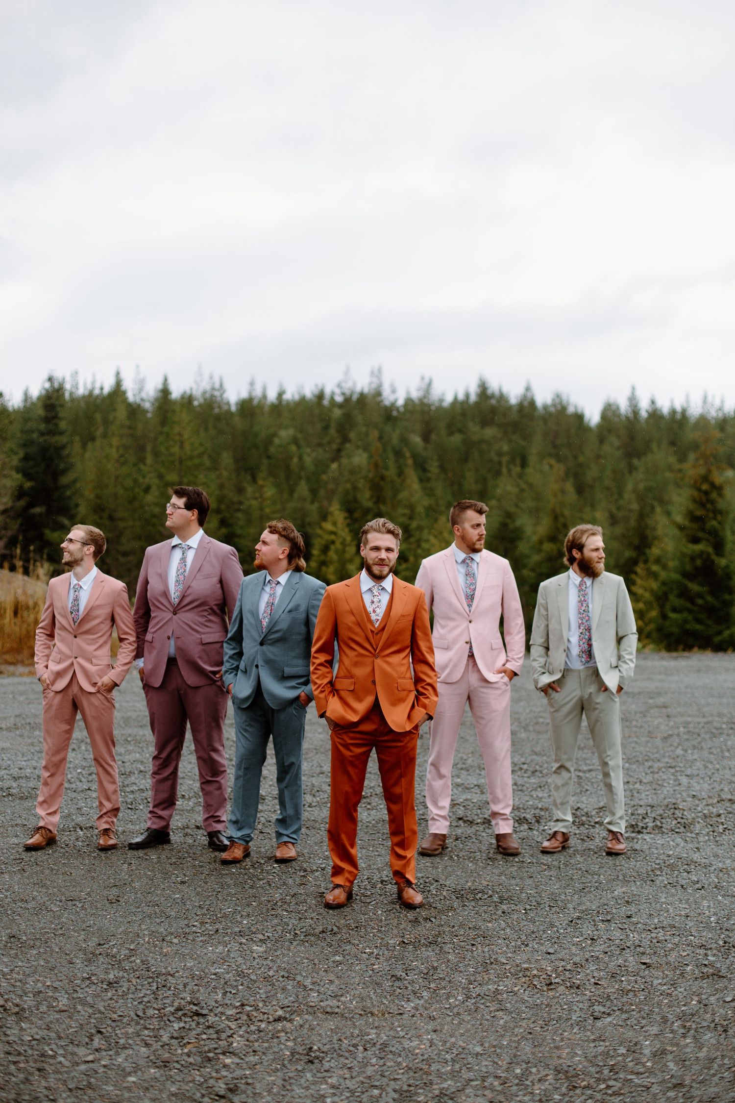 Six groomsmen in colorful mismatched suits pose outdoors on gravel with evergreen forest backdrop at a wedding.