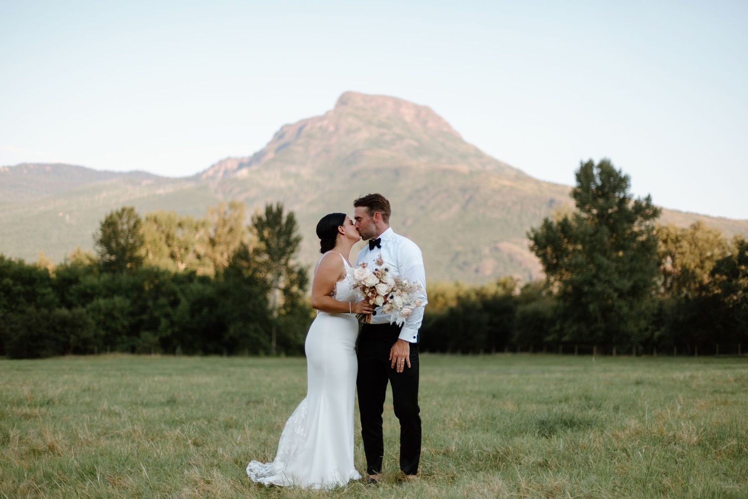 A couple shares a romantic kiss in a scenic mountain meadow at sunset with golden light illuminating the landscape.