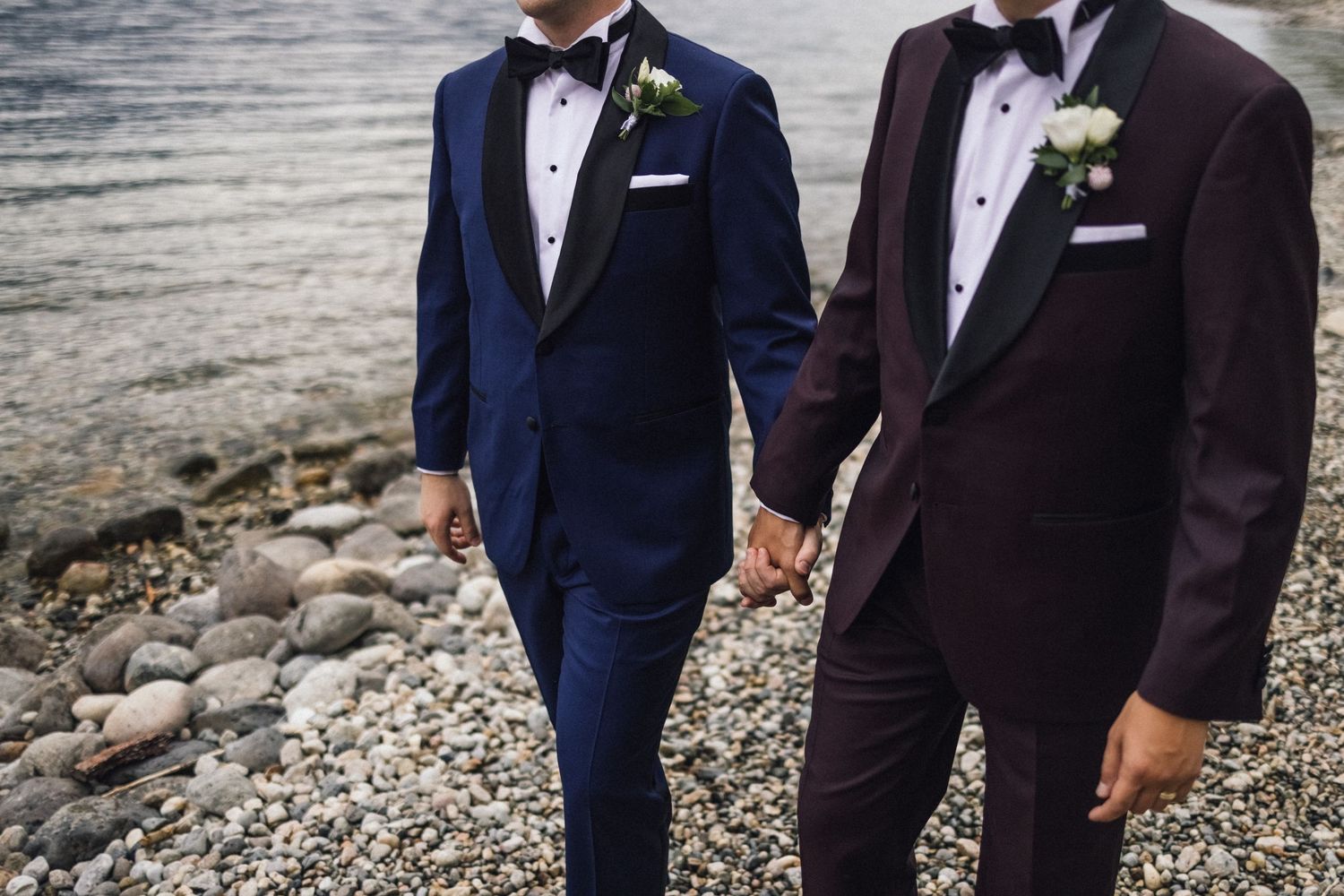 Two grooms in formal tuxedos hold hands on rocky beach with mountain backdrop.