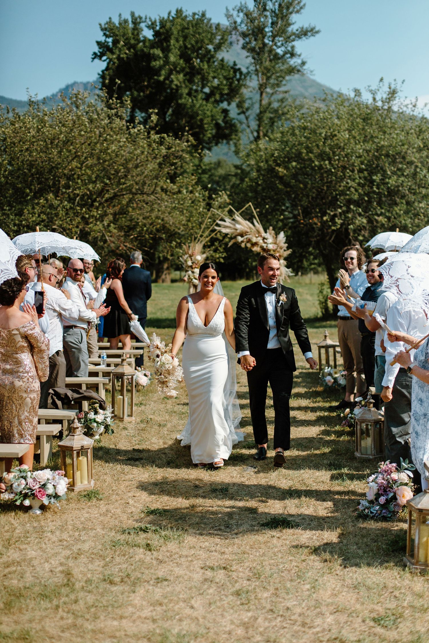 Guests celebrate at an outdoor wedding ceremony with rustic wooden benches set up in a grassy field with mountains in background.