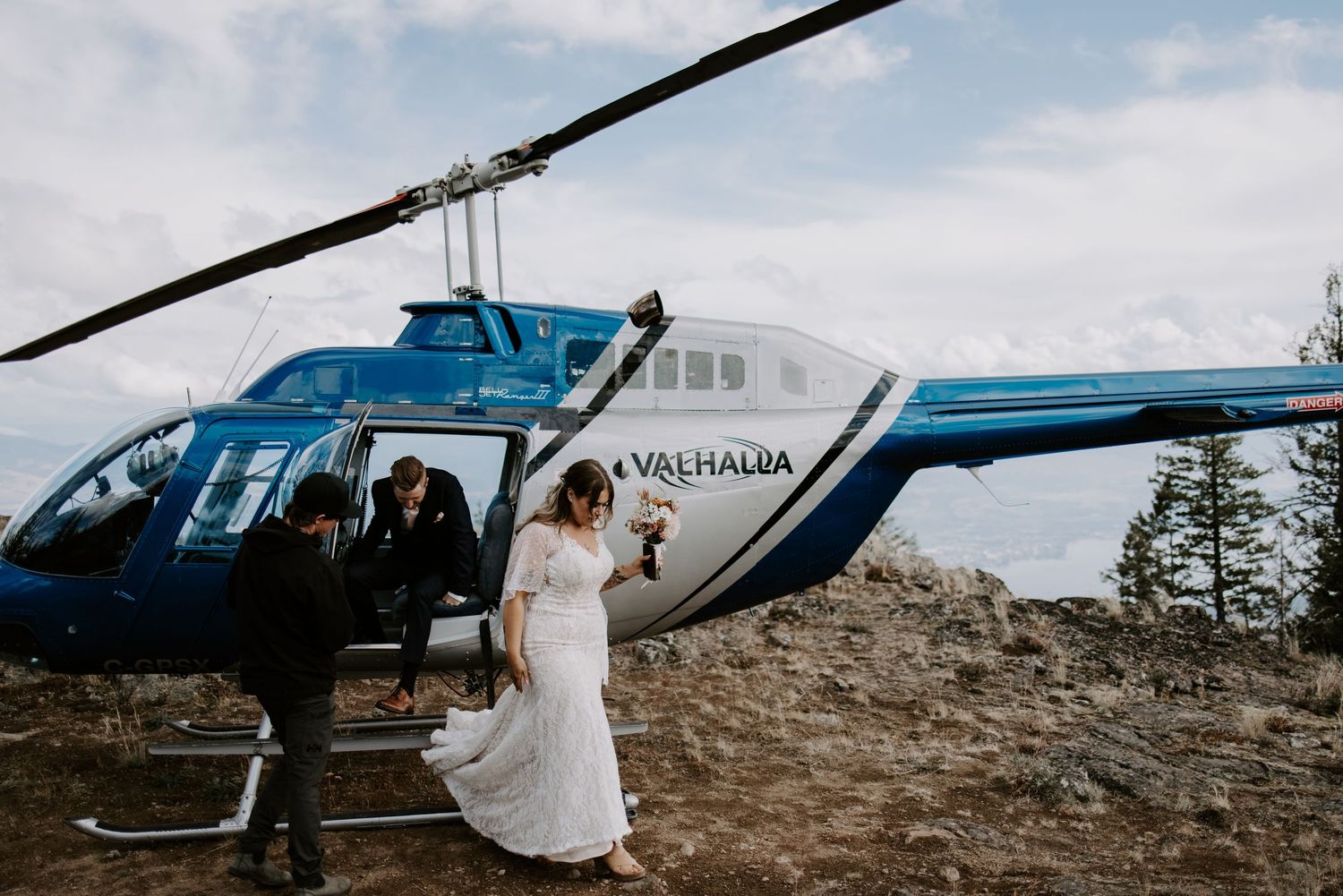 A bride in a white dress boards a blue helicopter on a mountainous terrain.