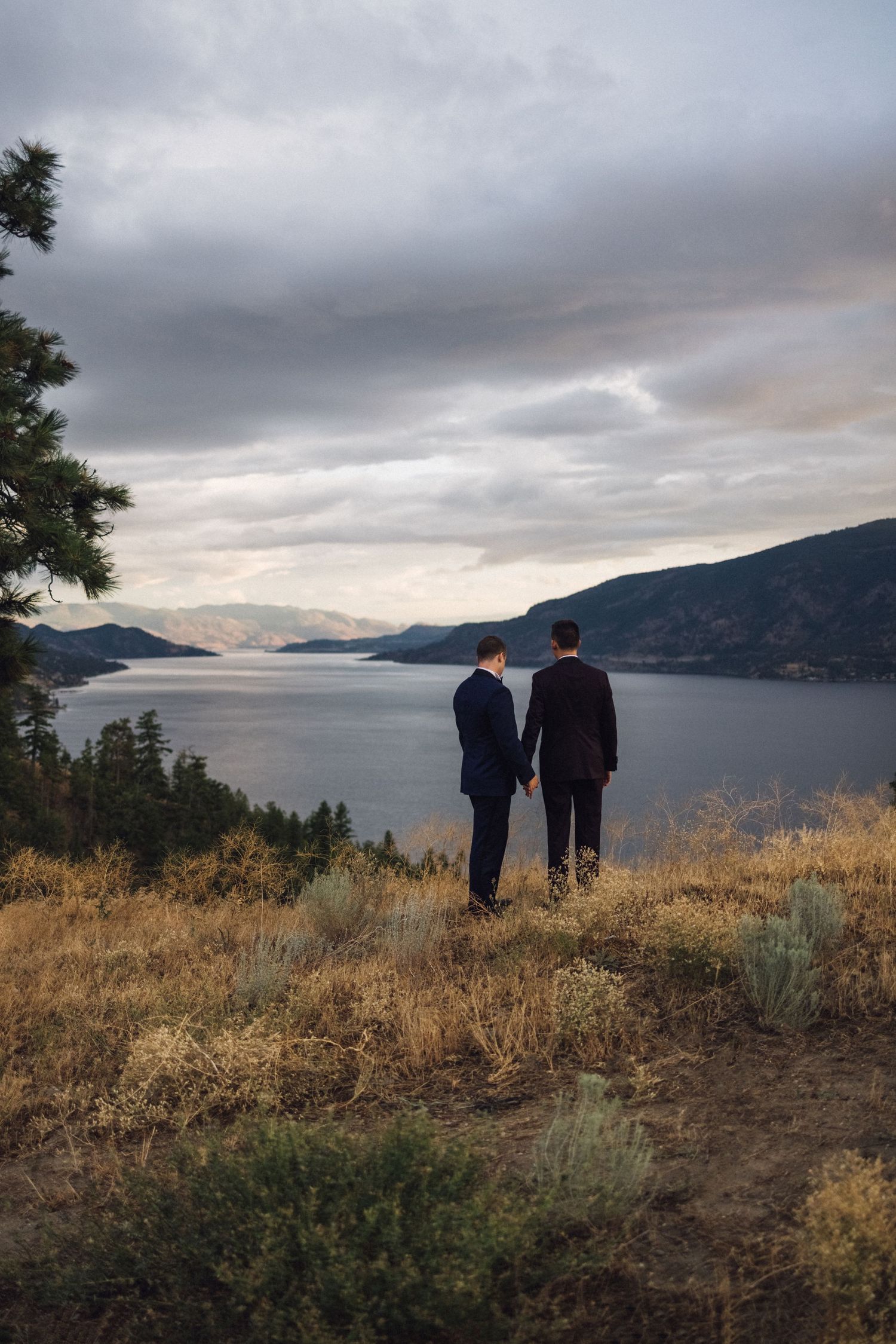 Two silhouettes stand against a dramatic mountain lake landscape at sunset, creating a peaceful scenic moment.