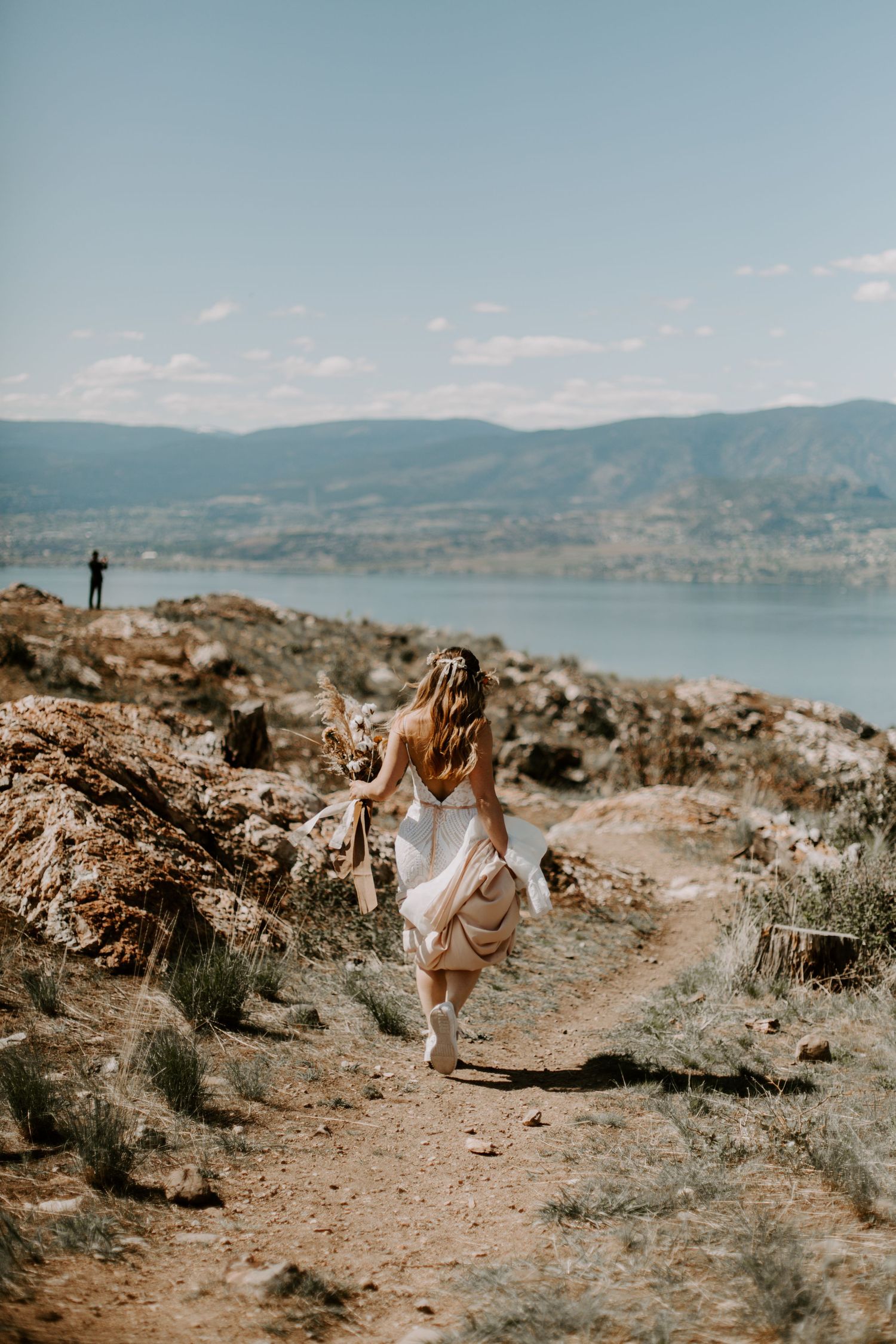 Person in flowing dress walks along rocky mountain trail overlooking scenic lake and mountains on sunny day.
