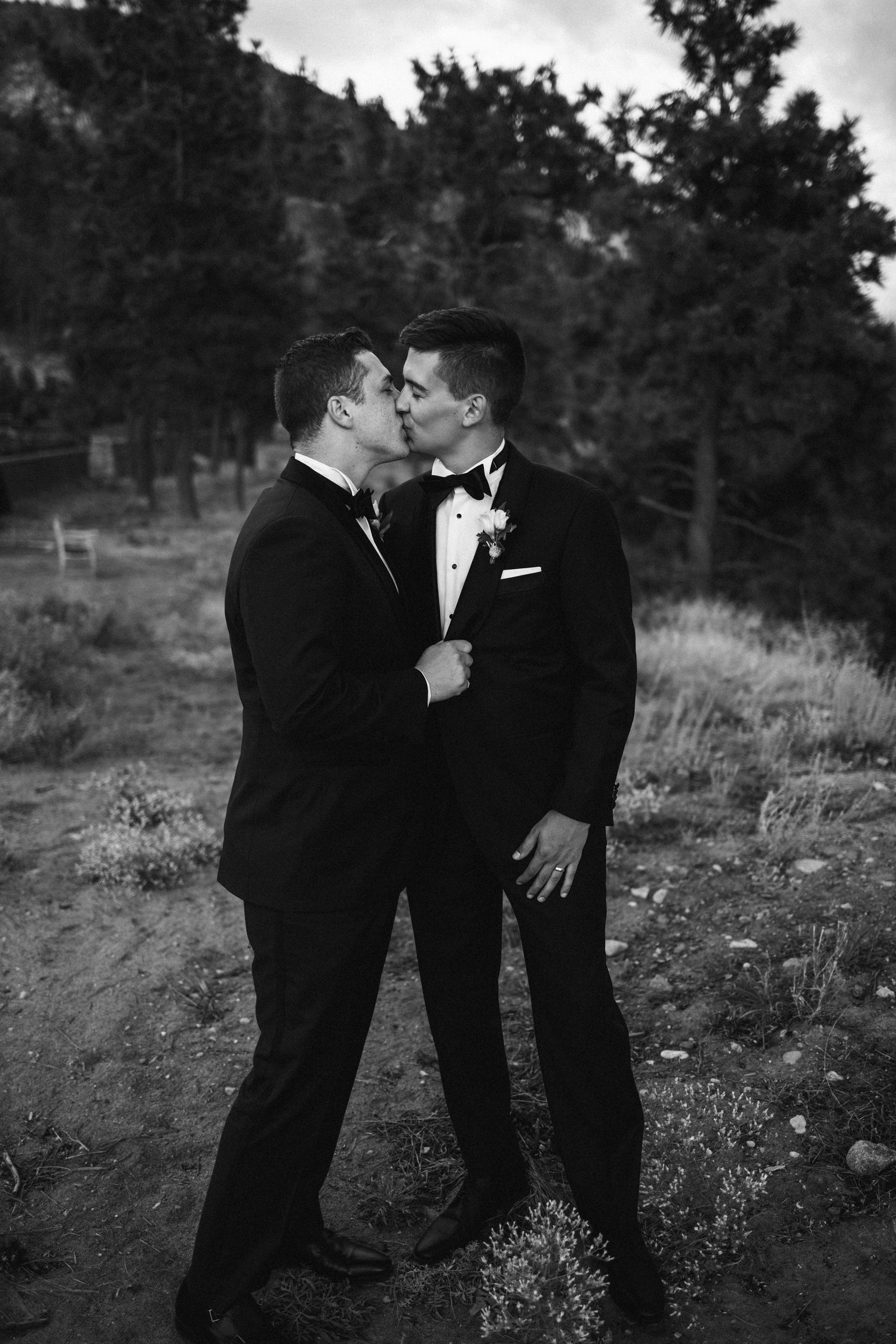 A black and white wedding photo of two grooms in tuxedos sharing an intimate moment outdoors.