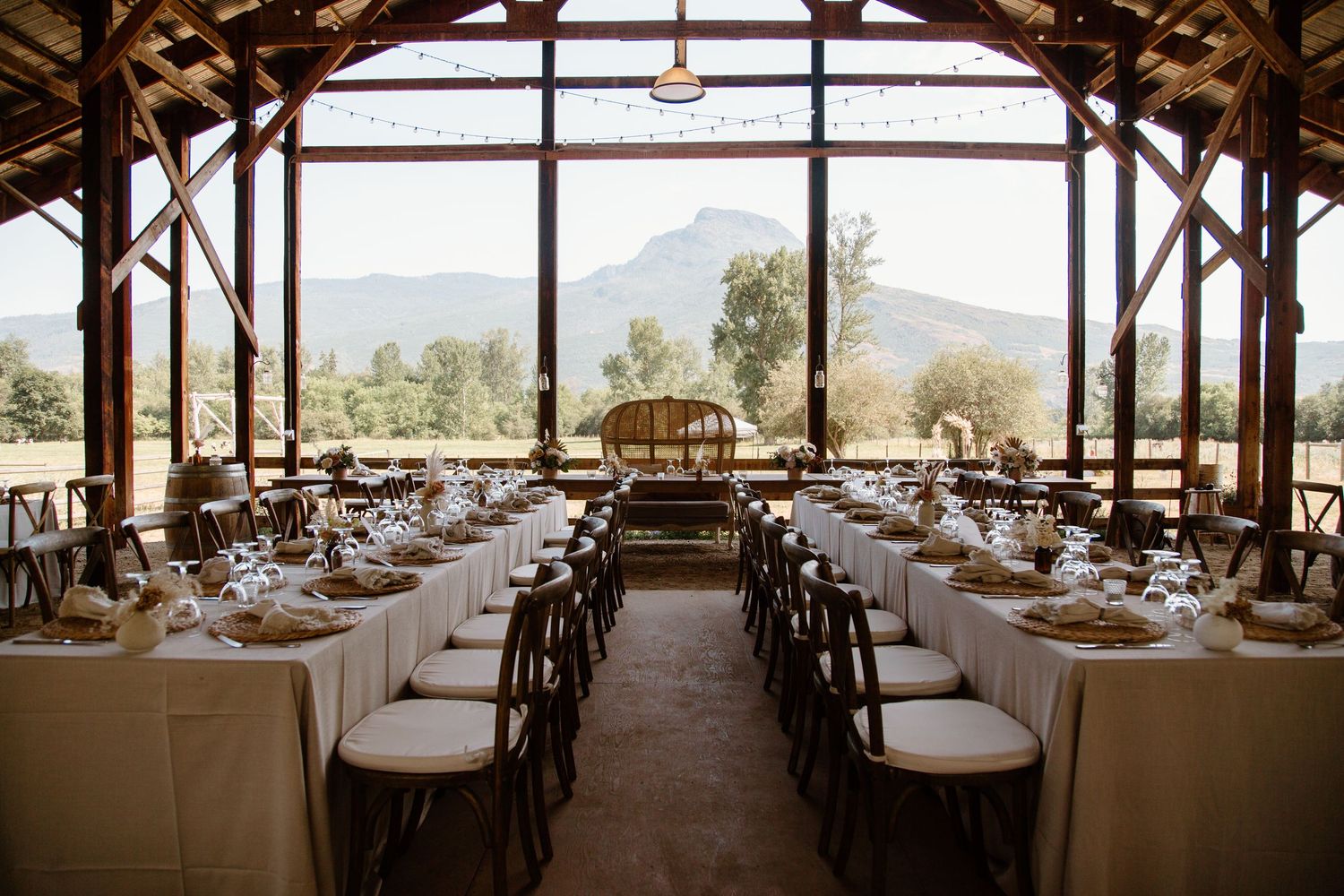 Elegant rustic wedding reception setup in a barn venue with mountain views through large windows.
