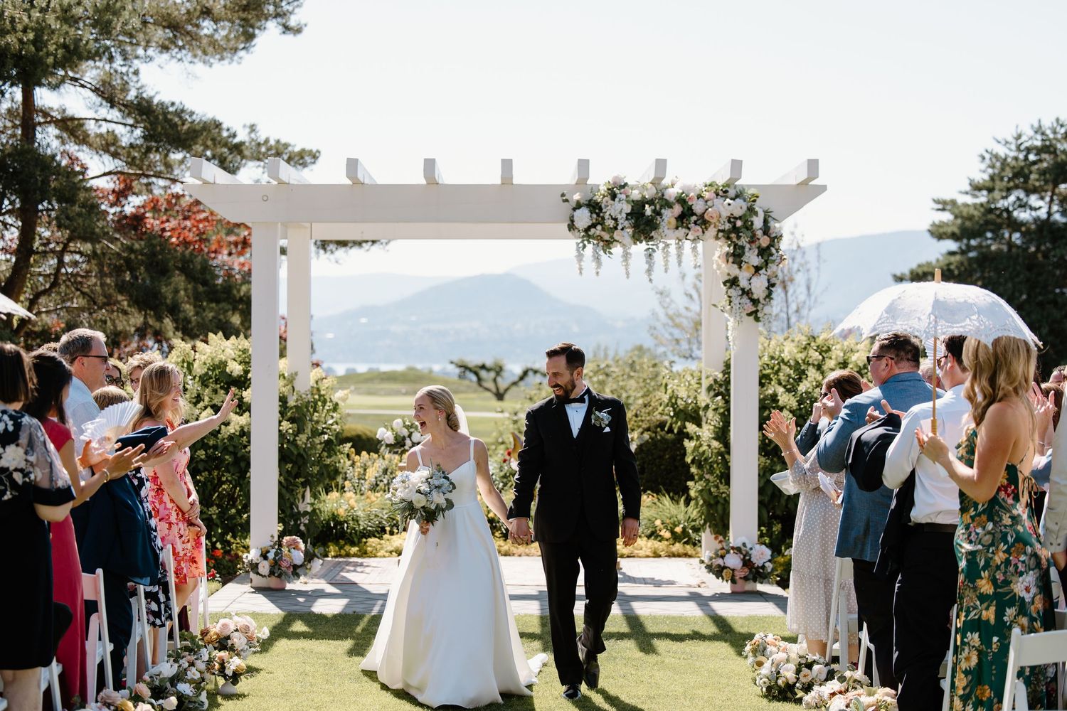 Newlyweds exit their ceremony under a white pergola as guests celebrate.