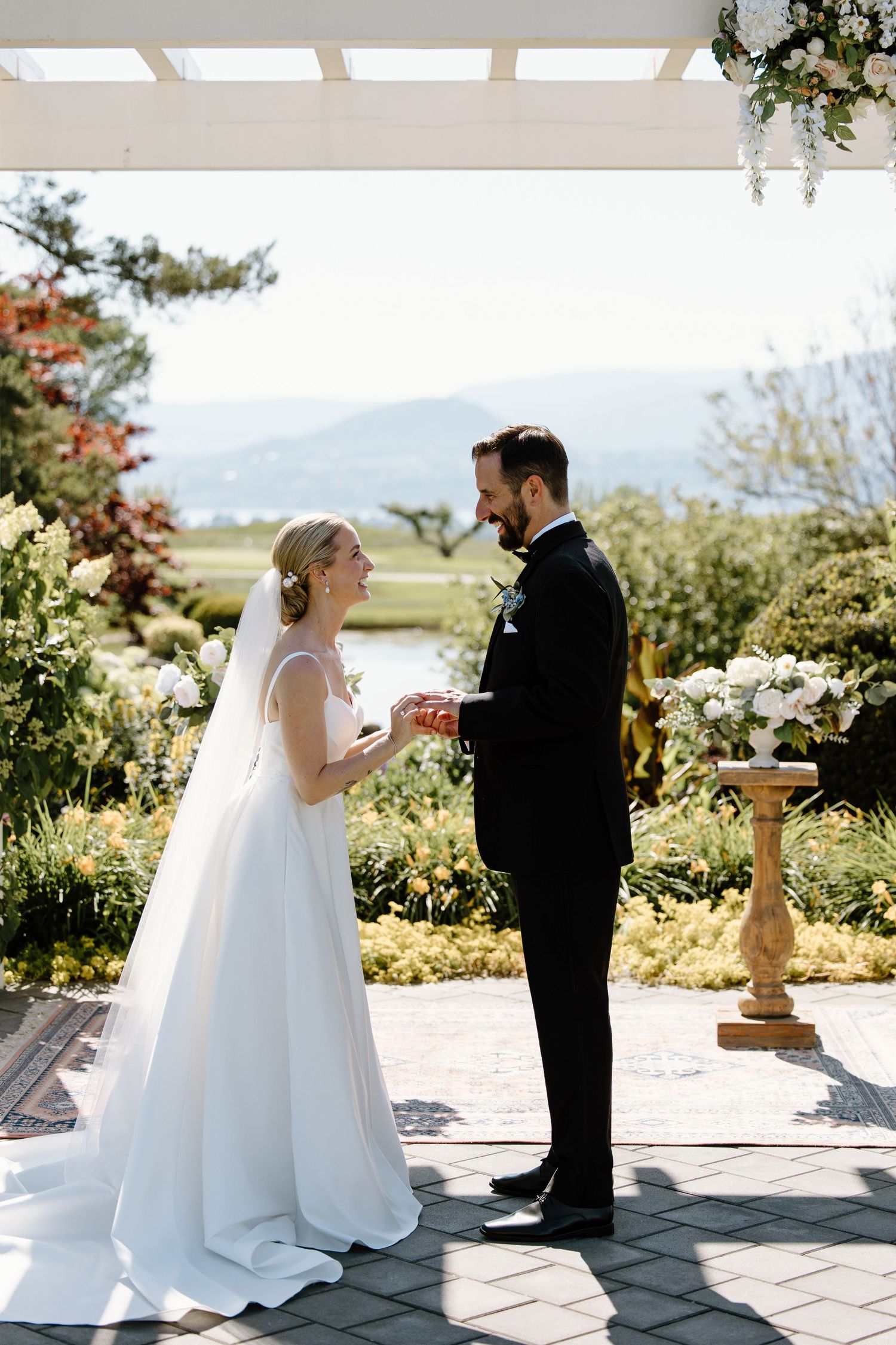 Couple exchanges vows under a white pergola with mountain views and garden flowers.