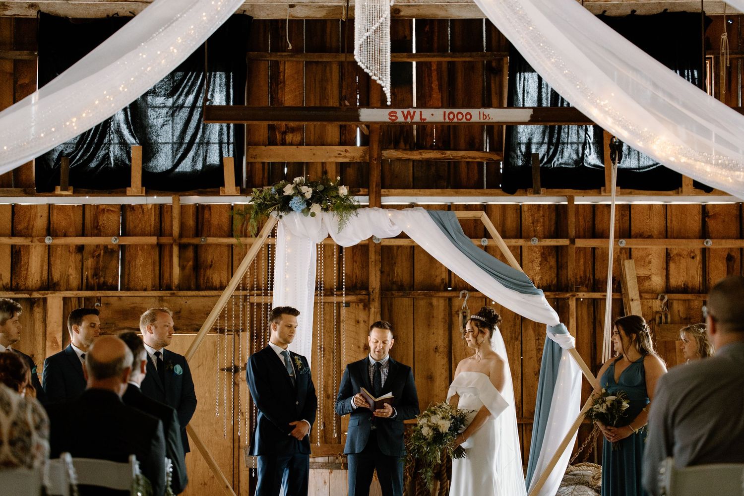 Rustic barn wedding ceremony with couple at floral arch, draped white fabric, and wooden walls.