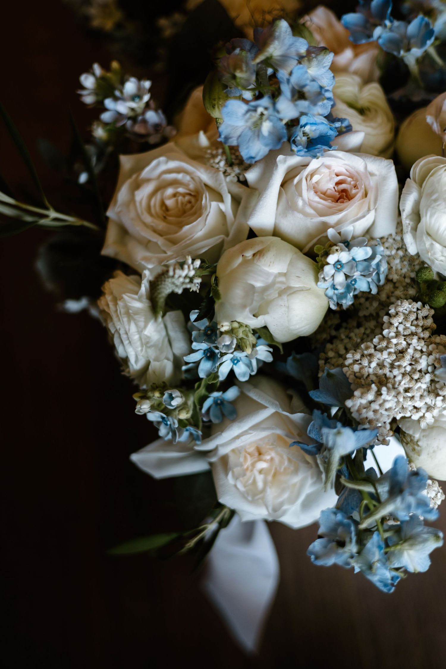 A romantic wedding bouquet featuring cream roses and light blue hydrangeas with delicate white accents against a dark background.