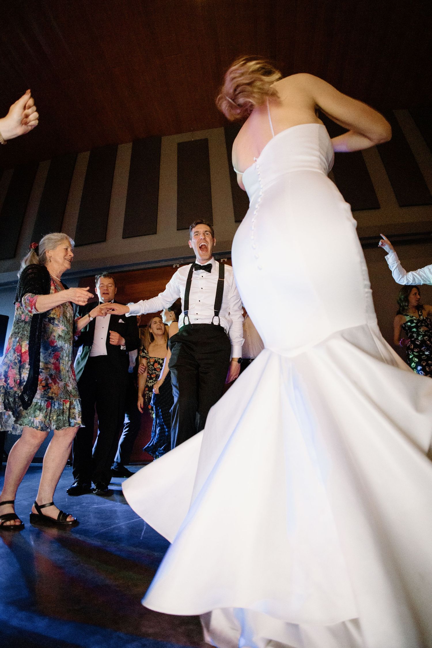 Bride in white mermaid gown dancing joyfully with groom in suspenders and wedding guests on reception dance floor.