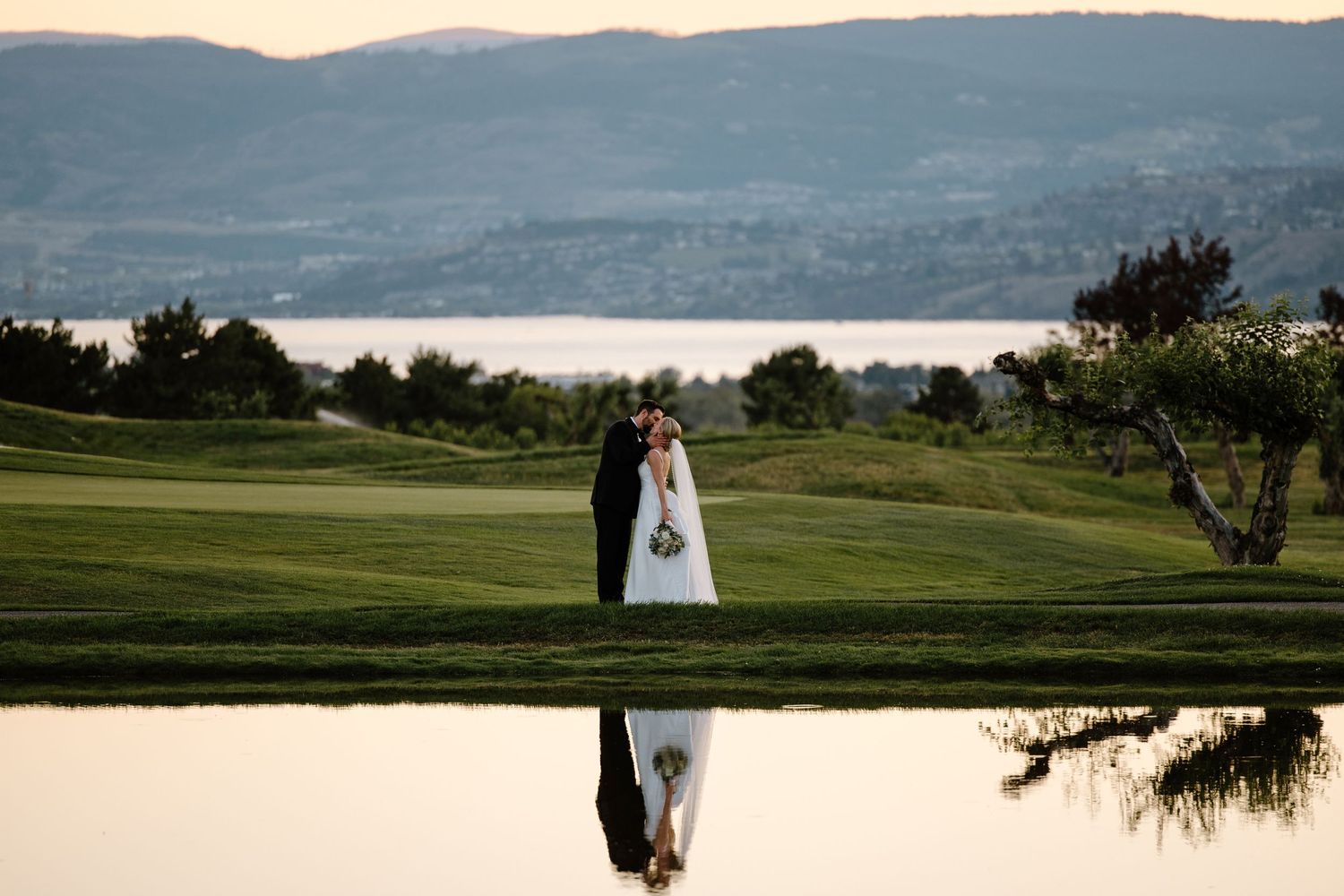 Bride and groom kissing on a lush golf course with a serene lake and mountain backdrop at sunset.