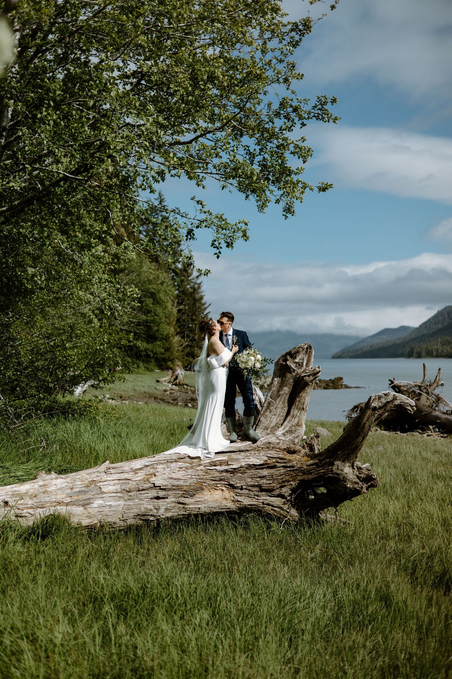 Bride and groom share a romantic kiss on a fallen log by a mountain lake, surrounded by lush greenery.