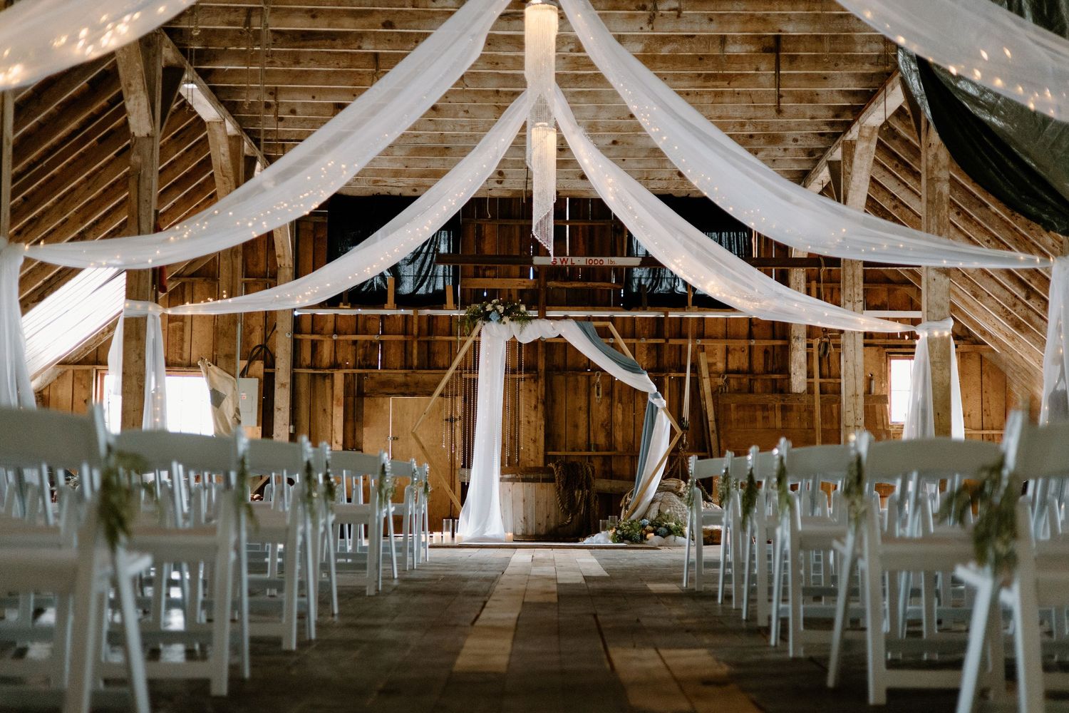 Rustic barn wedding venue with white draped ceiling, floral arch altar, and white chairs arranged in rows.