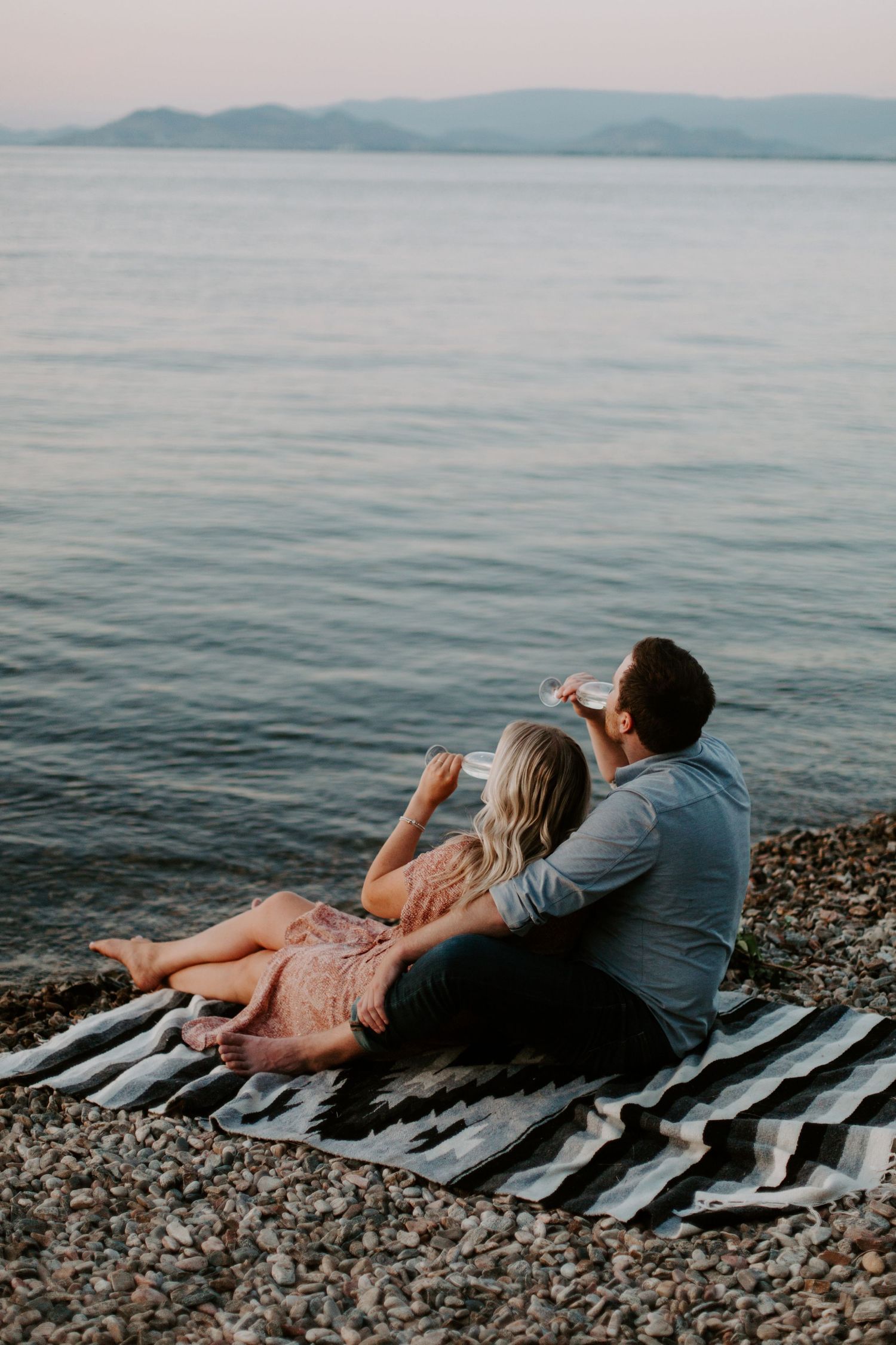 Couple sitting on striped blanket on rocky beach at dusk, drinking wine while enjoying calm ocean views.
