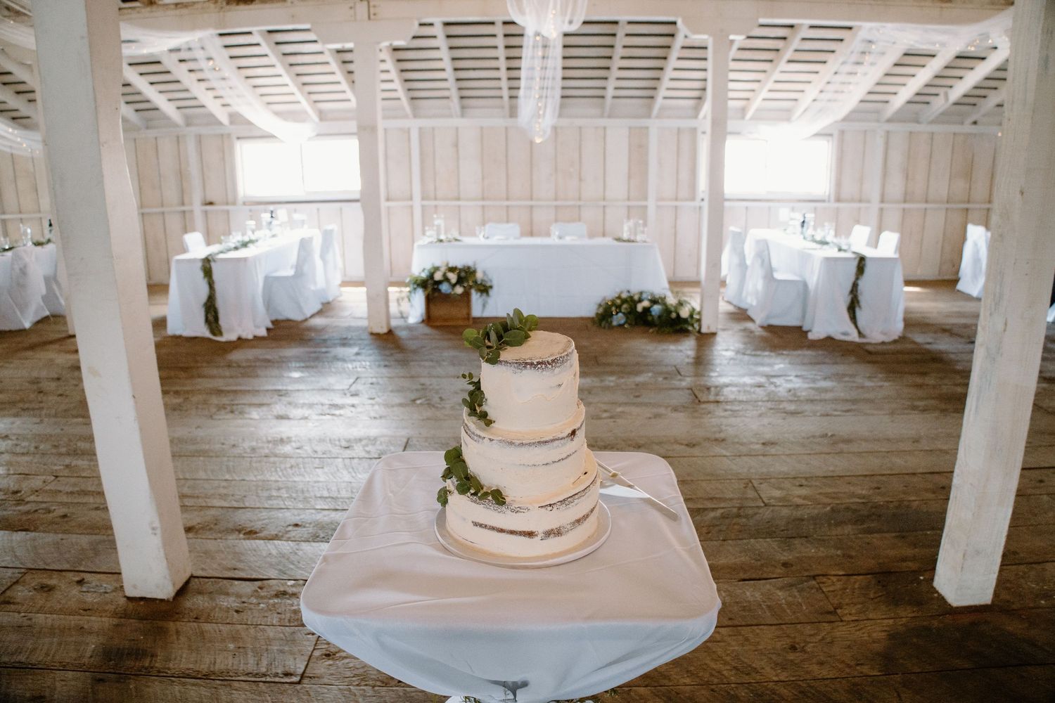 White wedding cake displayed on table in rustic venue with white columns and wooden flooring.