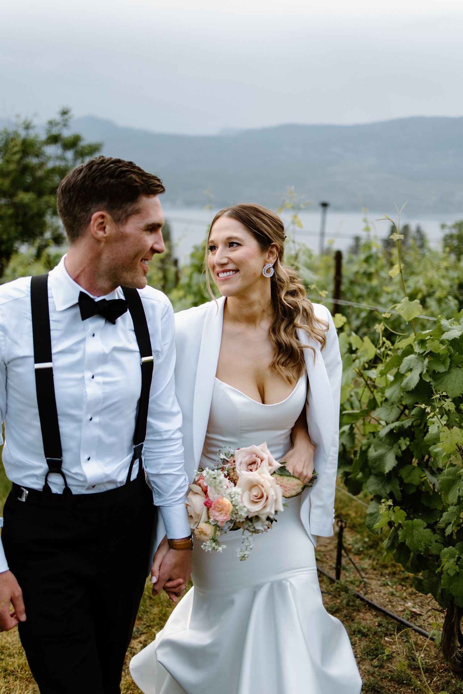 Newlyweds walking through vineyard with mountains in background.