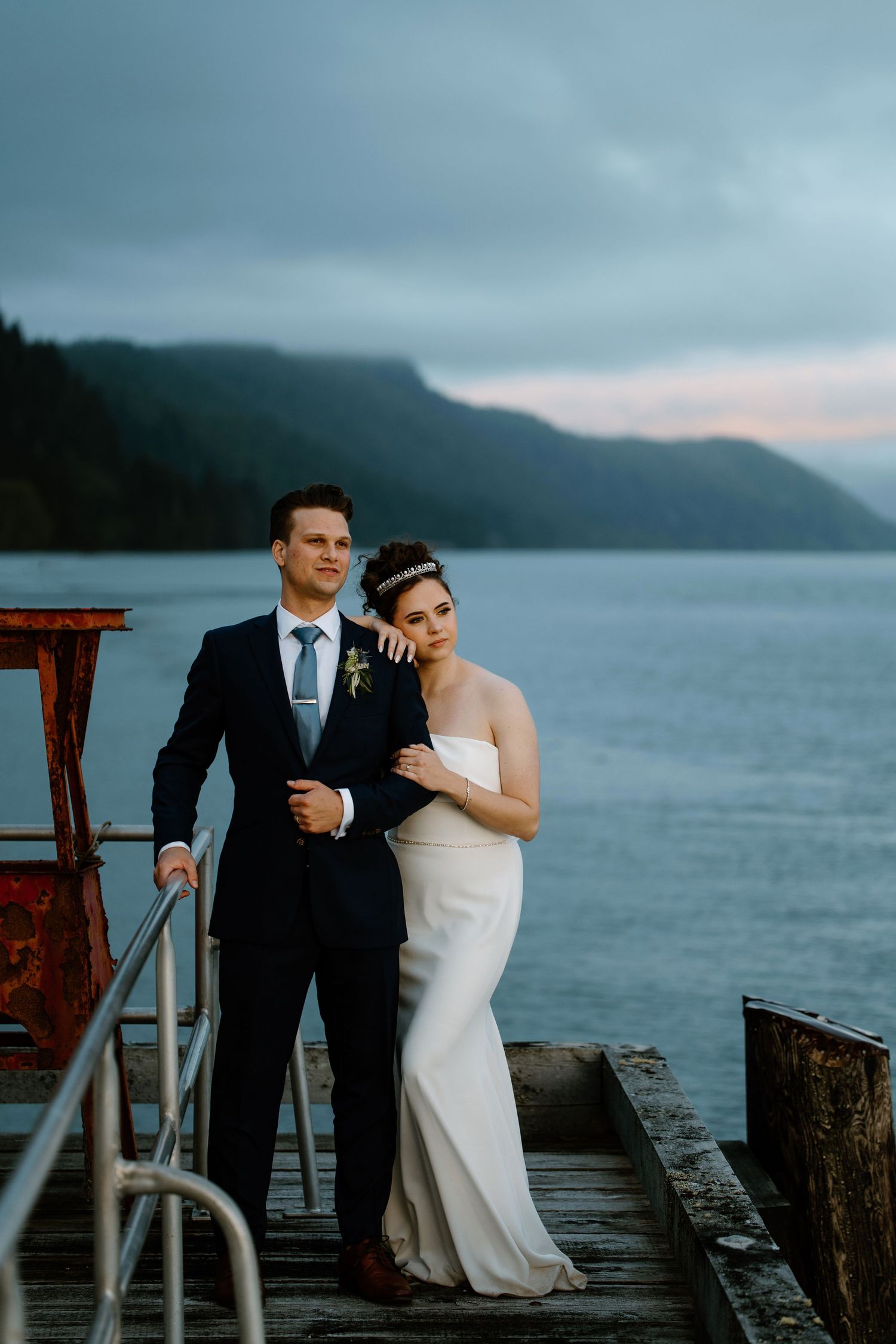 A romantic couple poses for wedding photos on a wooden dock by a misty lake with mountains in the background.