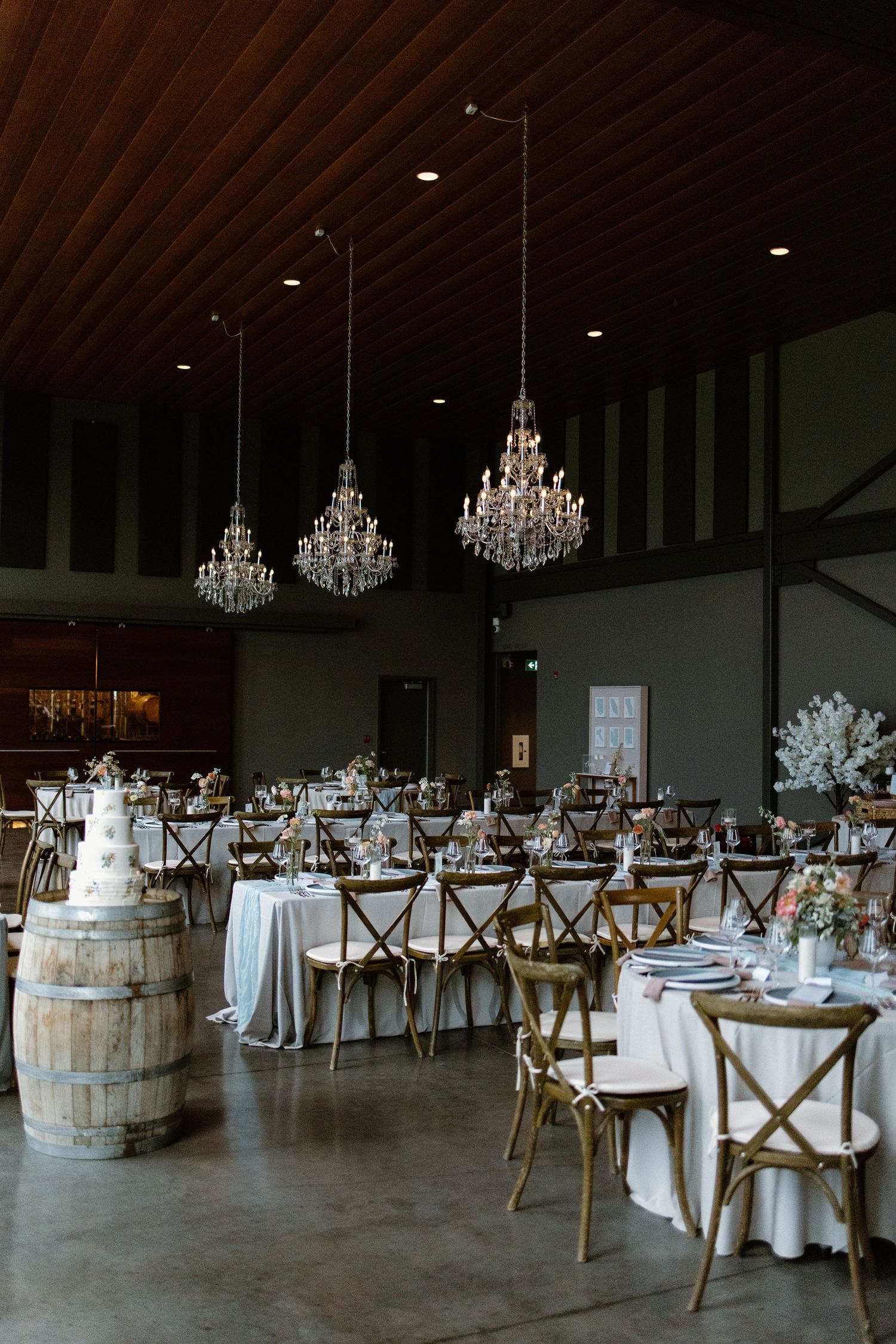 Rustic wedding reception space with wooden barrels, crystal chandeliers and cross-back chairs.