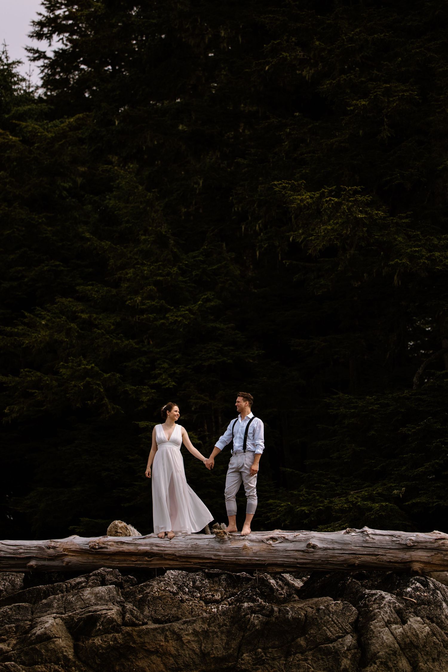 A couple in wedding attire sits together on a fallen log in a dark forest setting.
