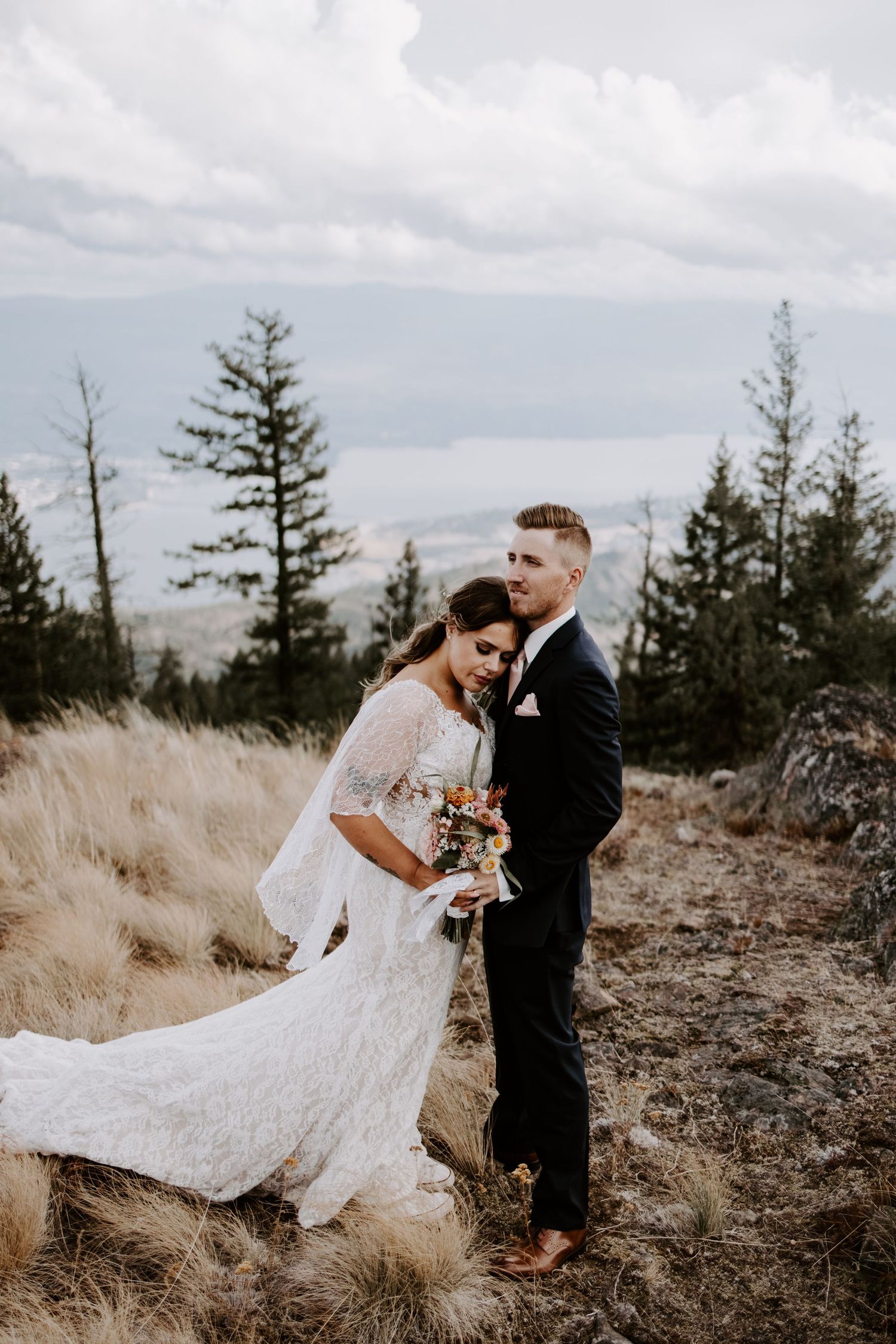 Wedding couple embraces on a mountaintop with evergreen trees and cloudy skies in the background.