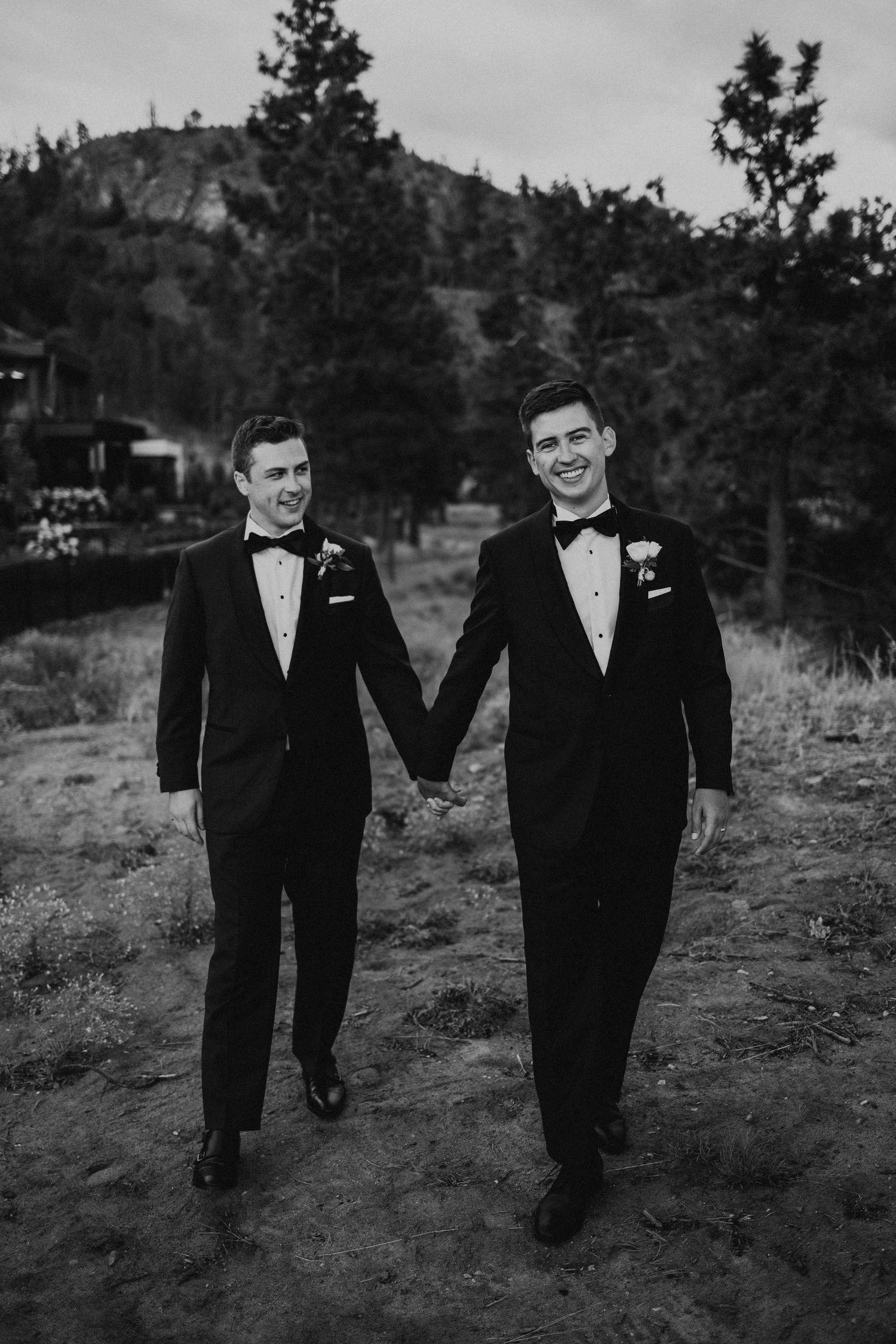 Two people in formal black tuxedos walking together outdoors in a black and white photograph.
