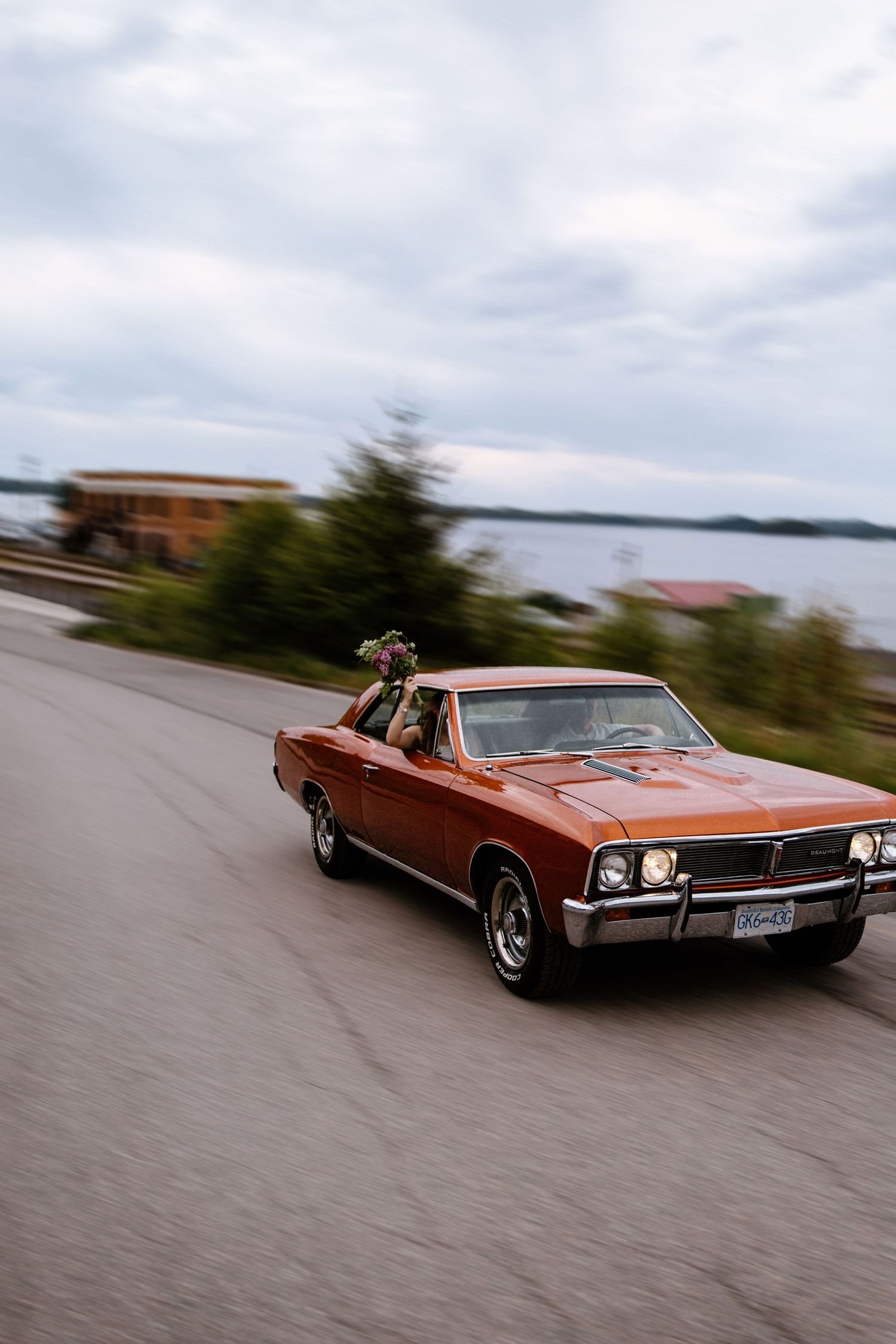 Classic orange Chevrolet El Camino driving on a coastal road with ocean and cloudy sky in background.