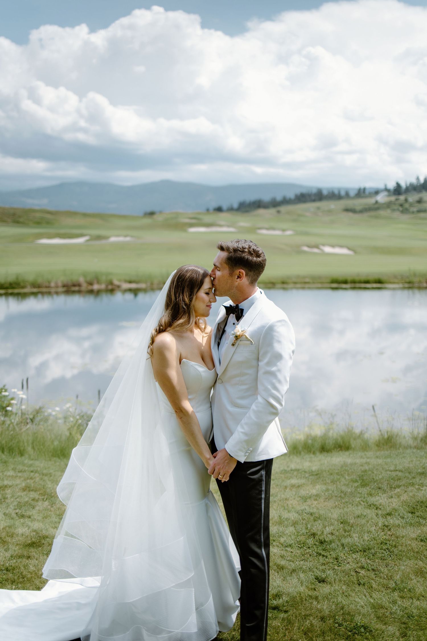 Bride and groom share a romantic moment by a mountain lake, she in a white gown, he in a white tuxedo jacket.