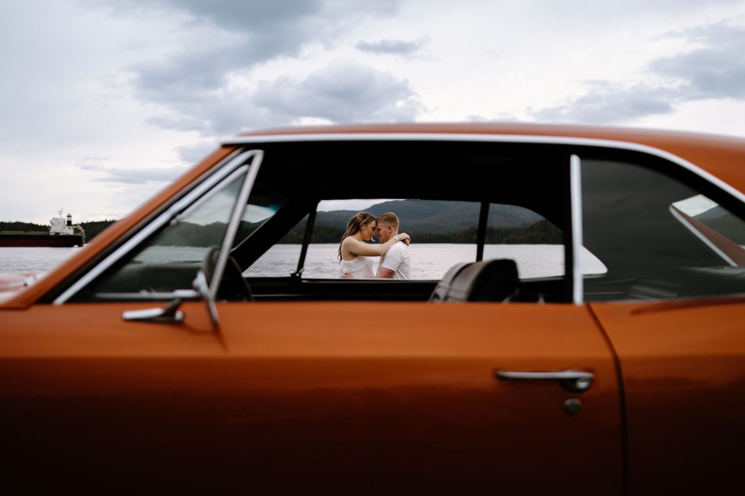 A couple sharing a romantic kiss inside a classic orange vintage car under a cloudy sky.