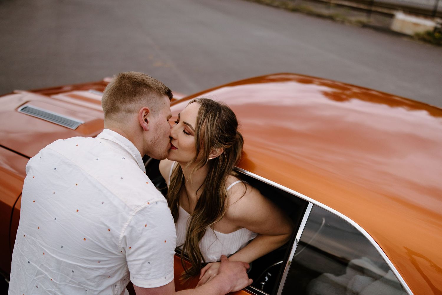 A romantic couple shares a tender kiss while sitting by an orange vintage car.
