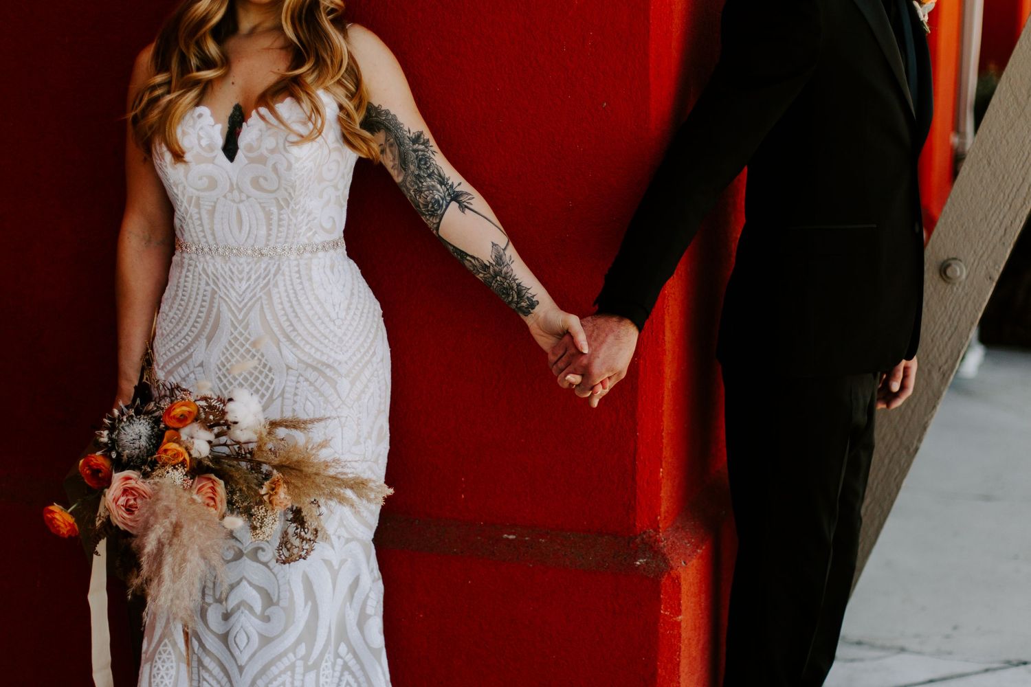 Detail shot of wedding couple holding hands against red wall, showing dress and floral bouquet.