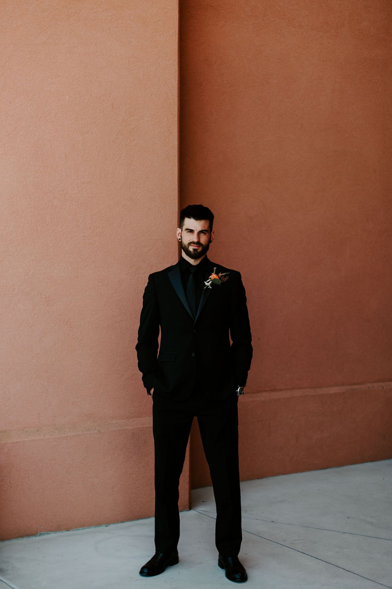 A person in a black suit stands against a terracotta-colored wall in a sequence of four similar poses.
